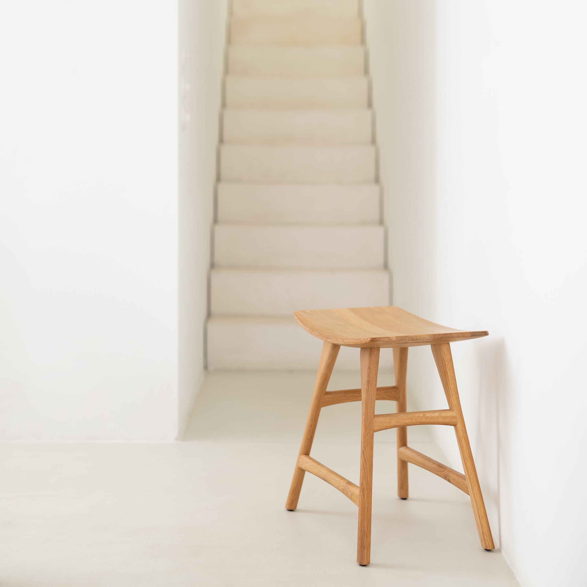 The Ethnicraft Osso Stool, crafted from oak or teak, is placed on a light floor in a minimalist hallway with a white staircase rising in the background.