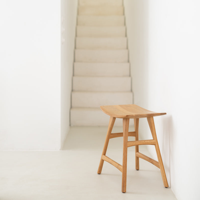 The Ethnicraft Osso Stool, crafted from oak or teak, is placed on a light floor in a minimalist hallway with a white staircase rising in the background.