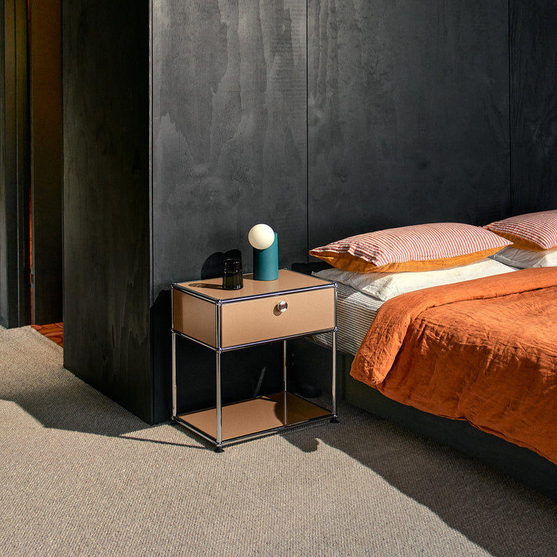 Haller Nightstand P2 in USM beige with chrome frame next to bed with orange bedding in modern bedroom.