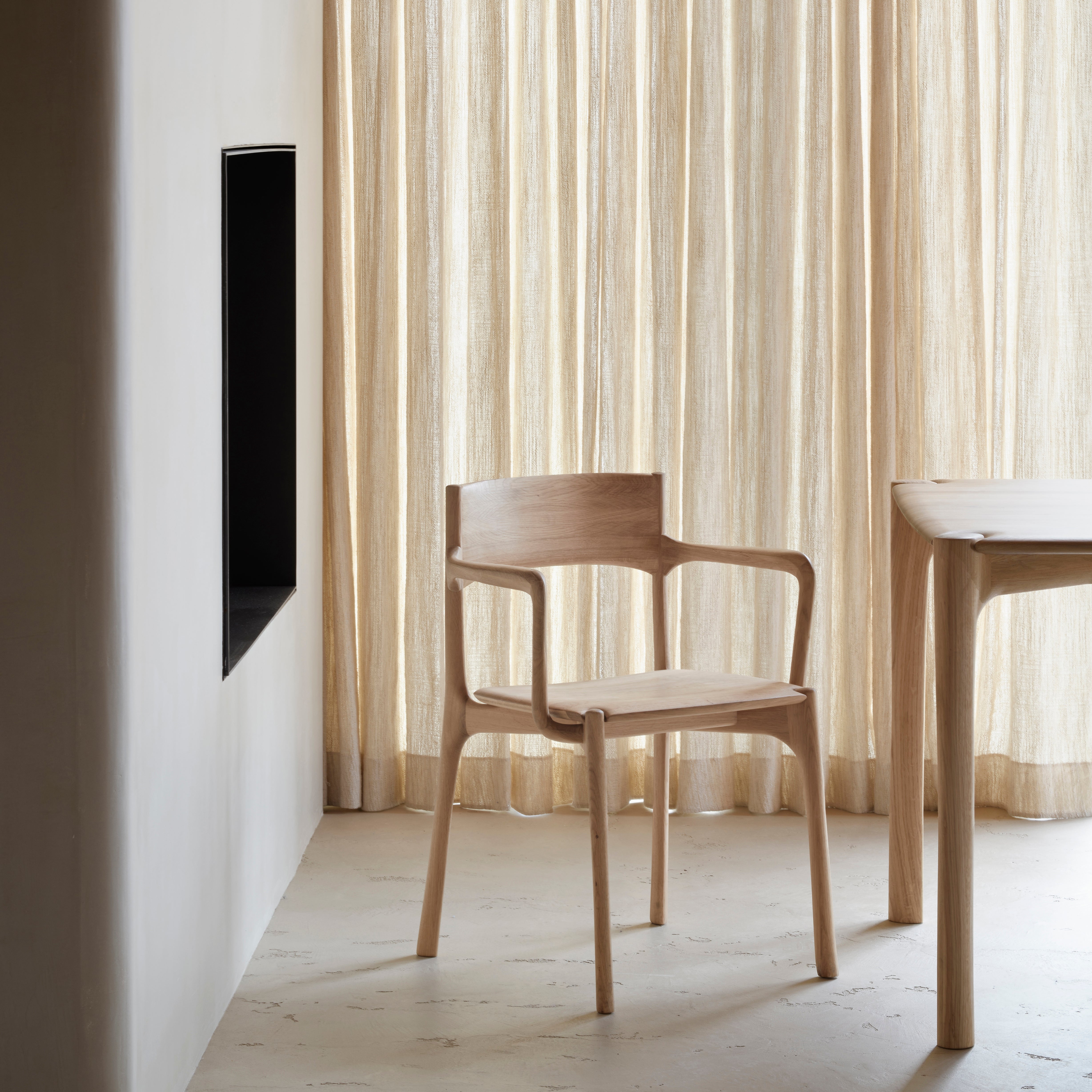 Pi Dining Chair with Armrest in oak wood featuring sleek, curved design and polished imperfect finish in a minimal dining room.