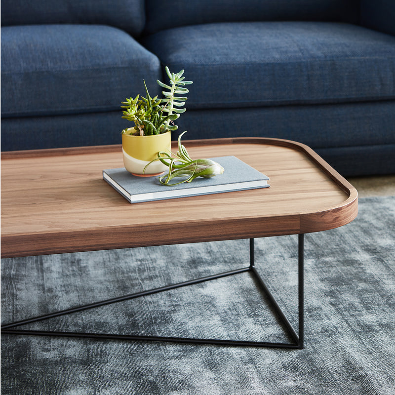 Rectangular walnut coffee table with raised rounded edges and slender black metal angular base on textured gray rug