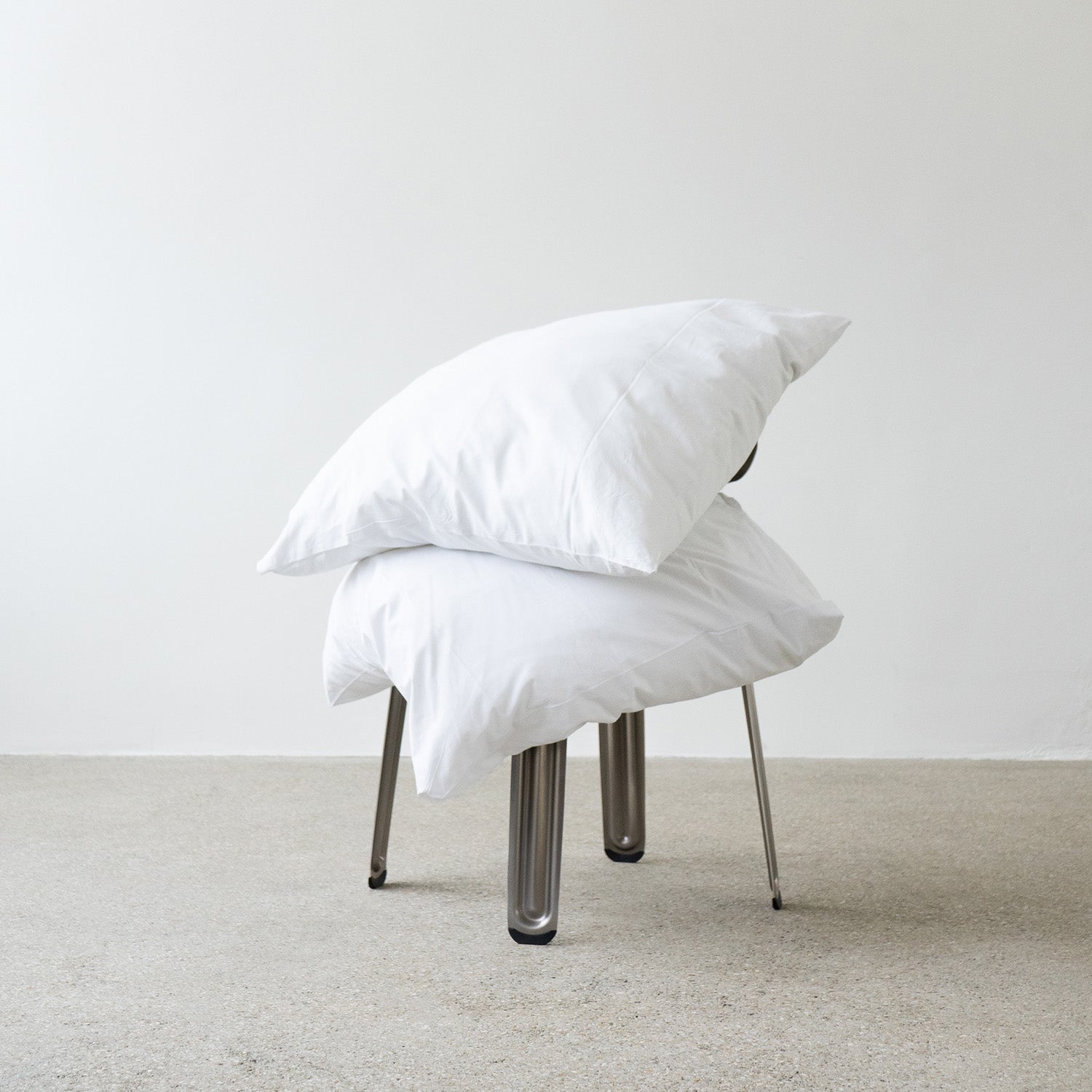 Two white pillows rest on a sleek Moormann Pressed Chair featuring metal legs, against a plain white wall and gray textured floor. The minimalist scene highlights the chairs stackable design.