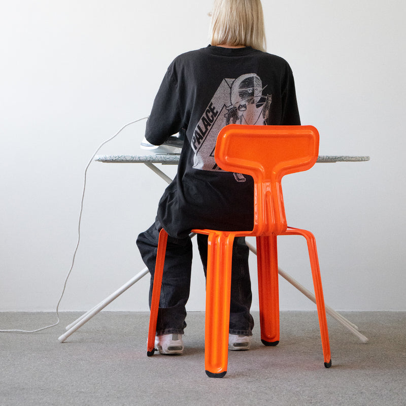 A person with long blonde hair sits on a bright orange Moormann Pressed Chair at a desk with a white surface and crossed metal legs. They wear a black graphic t-shirt and dark pants, set in a minimalistic space with gray carpet.