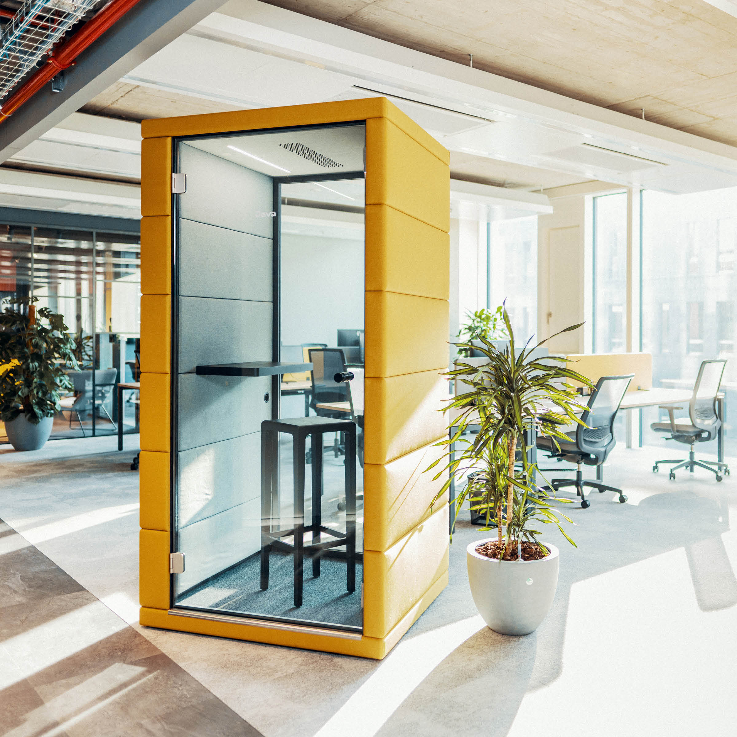 A modern office showcases a yellow phone booth with acoustic materials for noise reduction, featuring a stool and small shelf. Nearby, a potted plant thrives under bright window light. In the background, SilentLabs OFFICE POD PRIME integrates seamlessly with black chairs and desks.