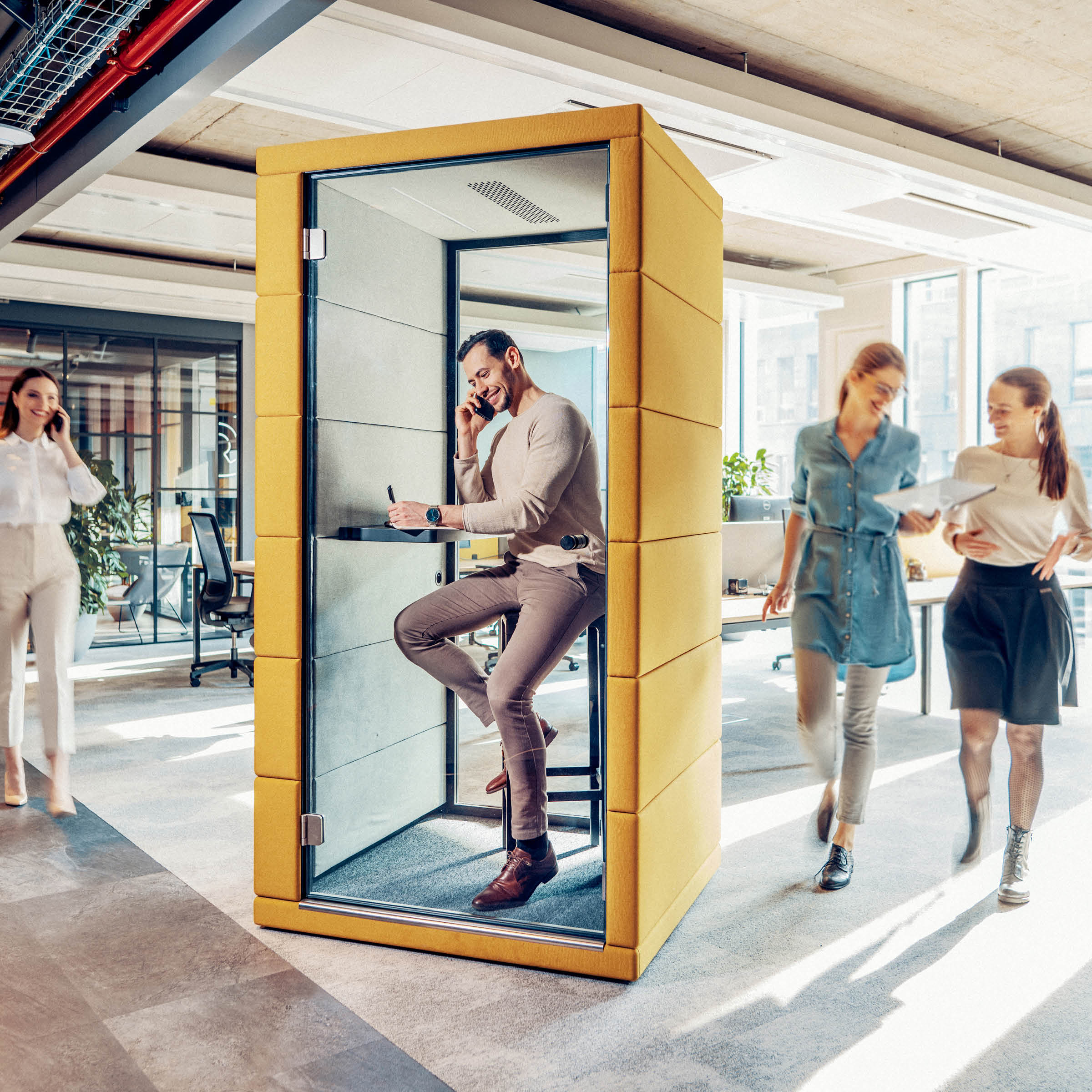 Inside a SilentLab OFFICE POD PRIME, featuring yellow acoustic materials for noise reduction, a man talks on the phone and writes in a notebook. Outside the soundproof booth, in the bright and modern office, three women pass by—two conversing and one on her phone.