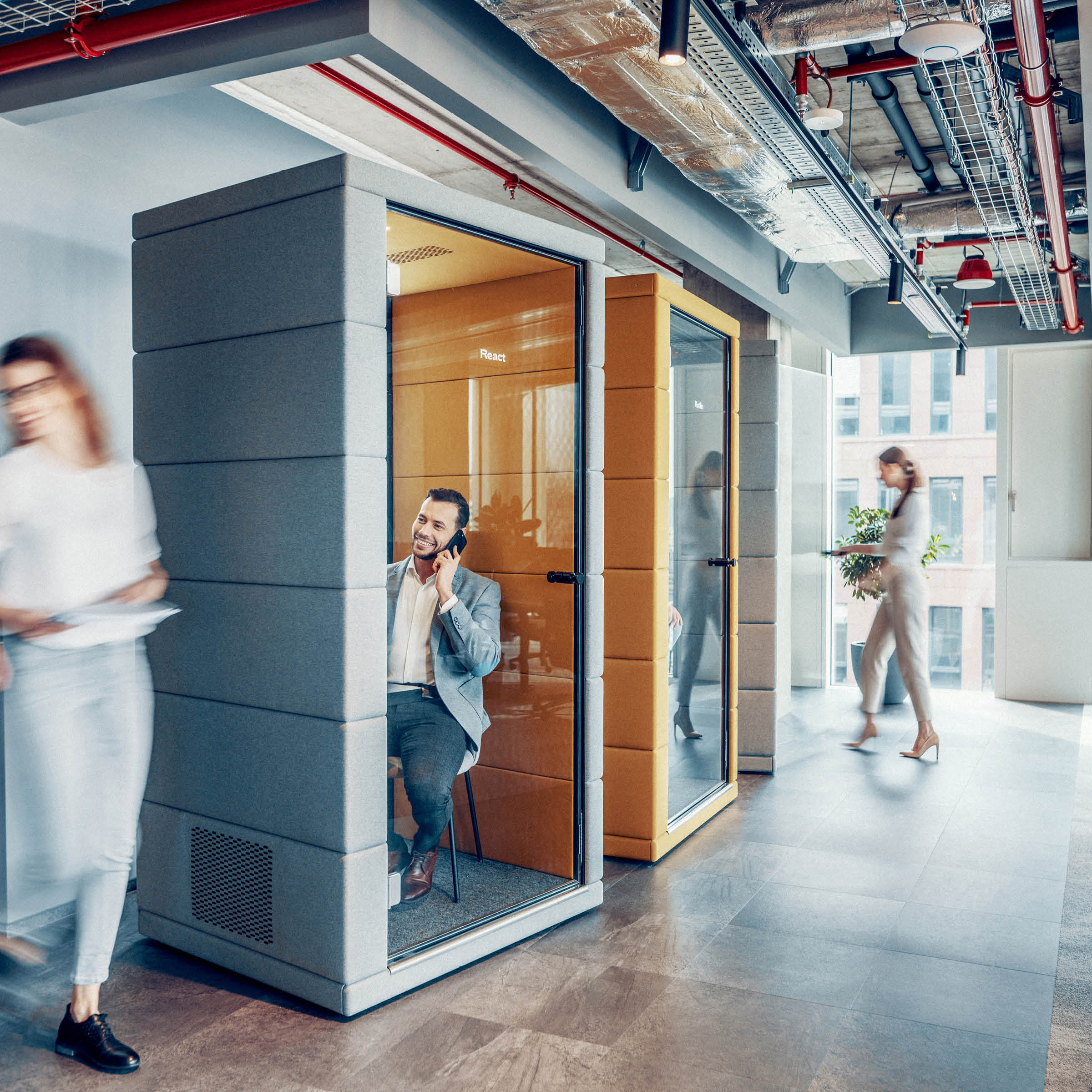 The office hallway includes SilentLabs OFFICE POD PRIME with noise reduction. A man inside a pod is talking and smiling, while two women walk past, one blurred. The area features industrial-style ceilings and large windows for natural light.