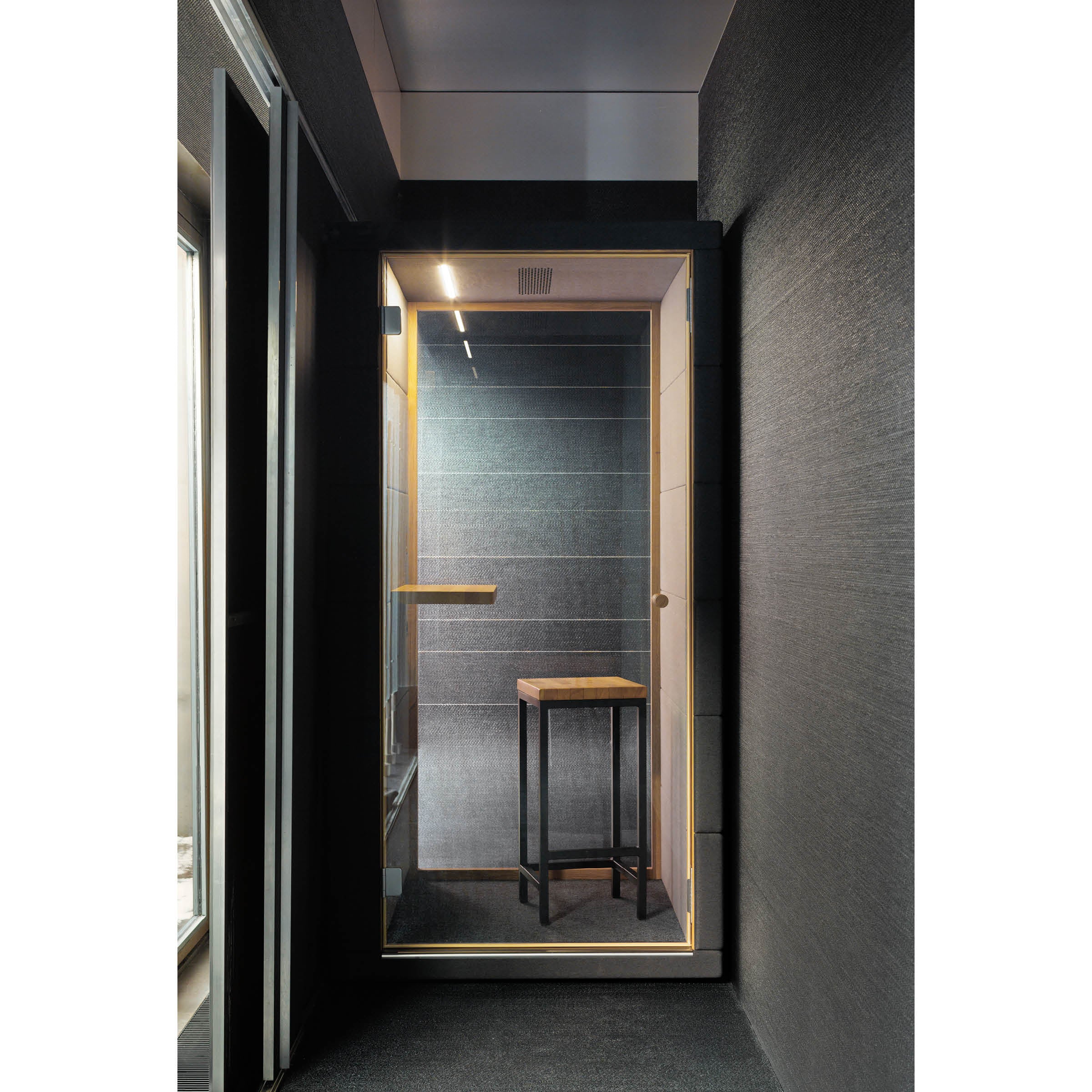 The OFFICE POD PRIME by SilentLab is a compact, modern soundproof booth with advanced acoustic materials. It features a wooden stool, wall-mounted shelf, glass door paired with dark acoustic panels, and a ceiling light for a cozy atmosphere.