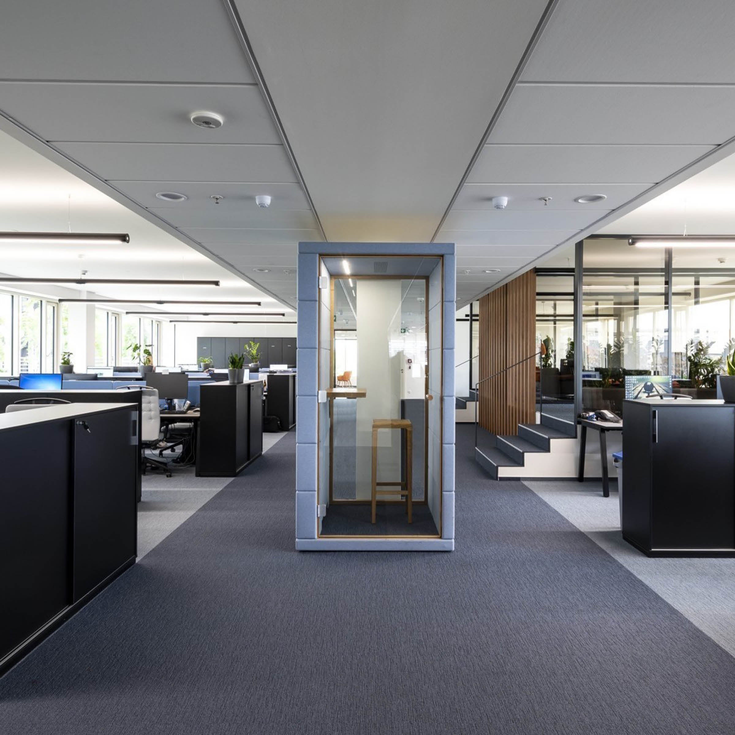A modern office space highlights the SilentLab OFFICE POD PRIME at its center, engineered for noise reduction. Its encircled by desks with black chairs, large windows, and glass conference rooms. The carpet is a blend of blue and gray tones, with plants distributed throughout.