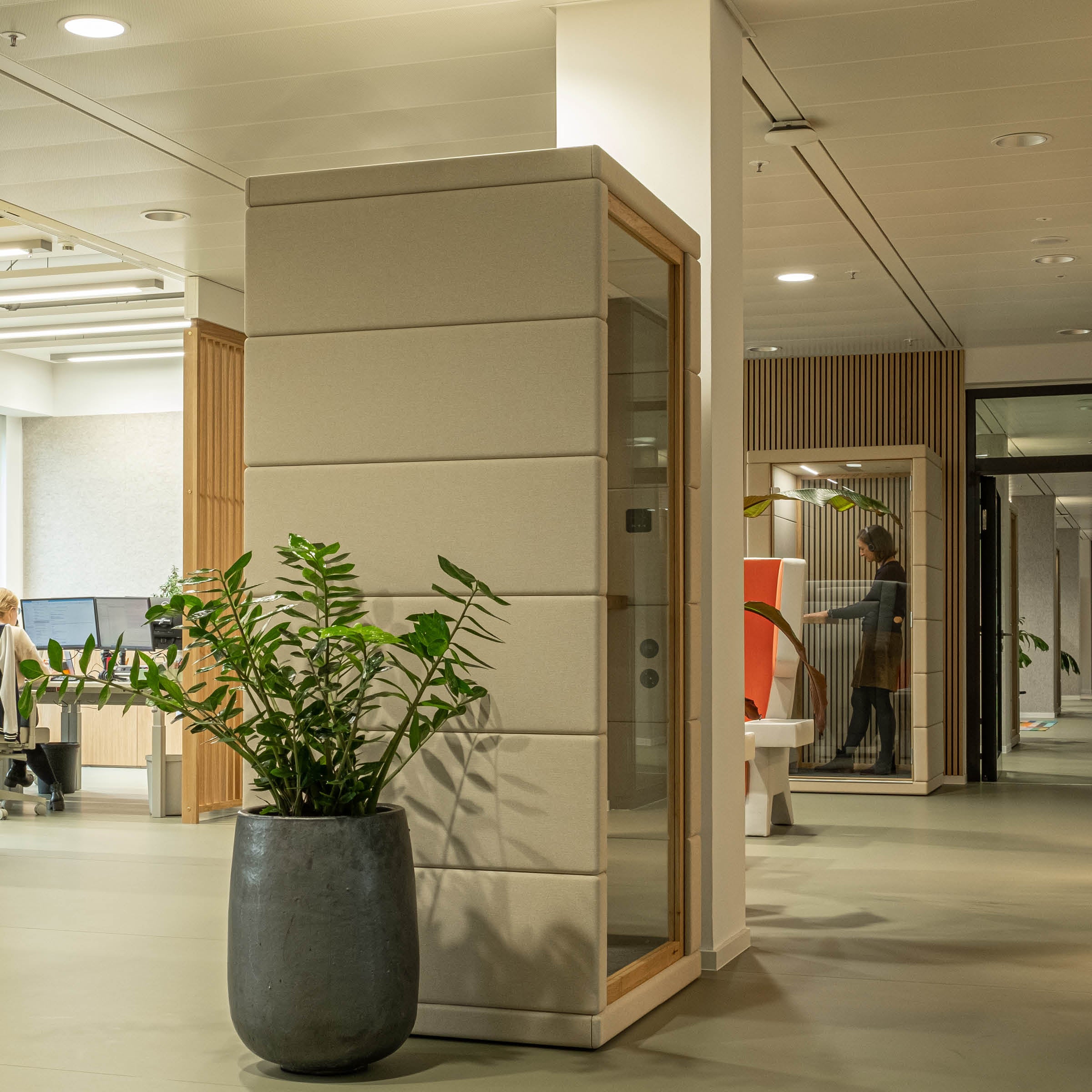 Office space with SilentLabs OFFICE POD PRIME, featuring modern soundproof phone booths made from cutting-edge acoustic materials. A large potted plant adorns the foreground, while open-plan workstations with natural light and wood accents create a cozy atmosphere, with a blurred person in the background.