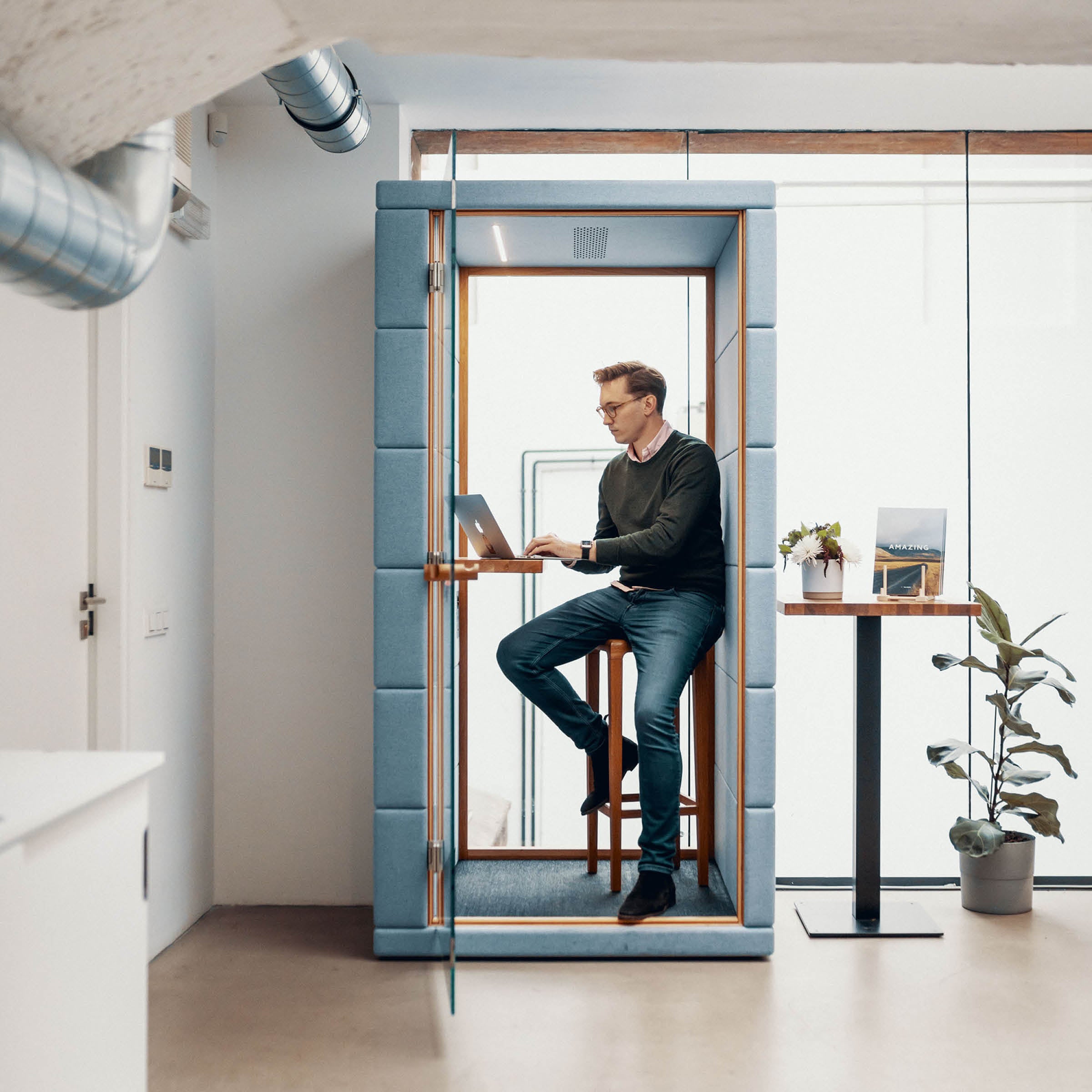 Inside a modern office, a casually dressed individual sits on a tall stool in the SilentLab OFFICE POD PRIME, working on their laptop. The small pod features glass walls and is surrounded by plants, a table, and acoustic materials for optimal noise reduction.