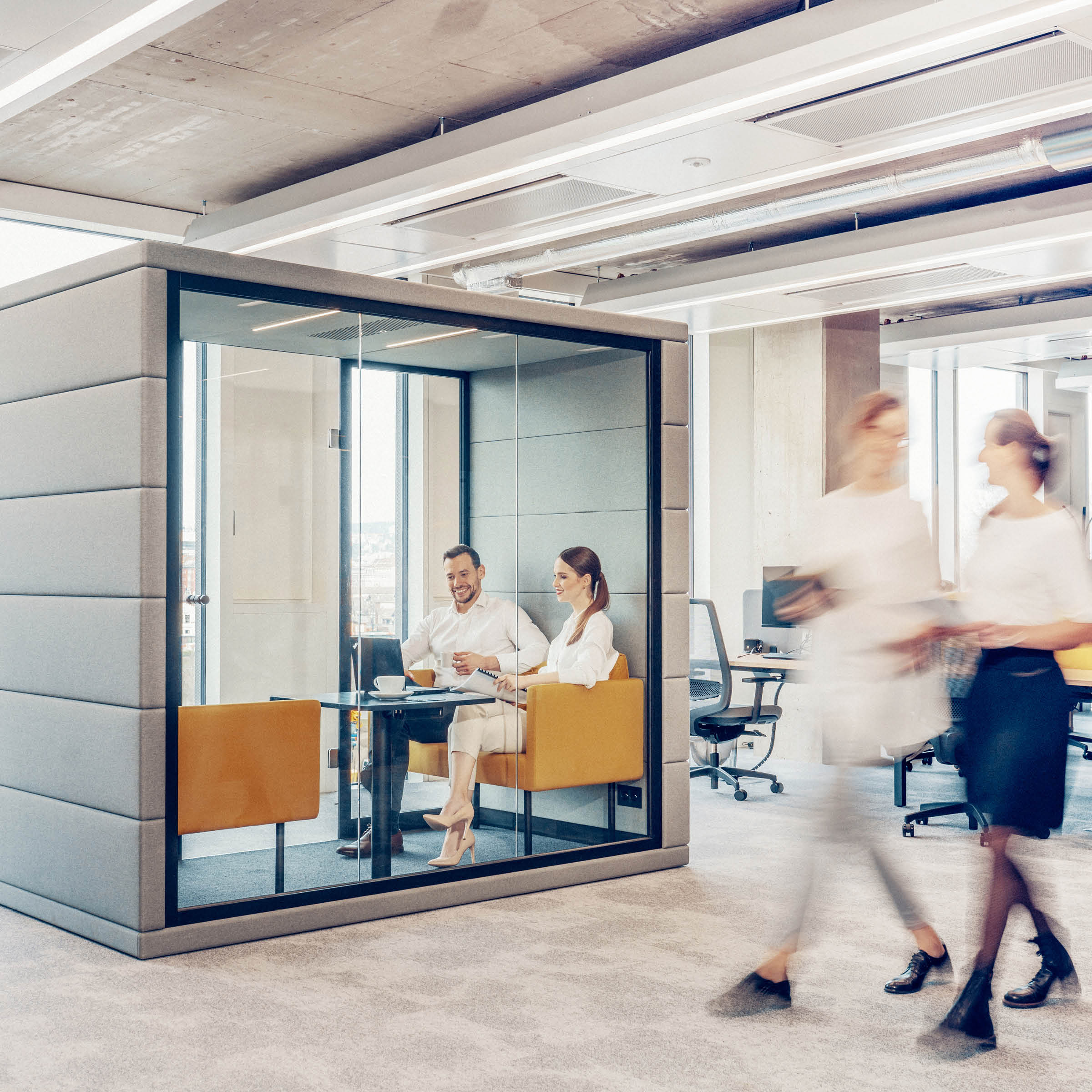 A man and a woman engage in conversation inside the modern SilentLab OFFICE POD QUADRIO 1, set in an open office. The pod’s bright lighting and neutral tones provide privacy and a contemporary design, as two blurred figures pass by outside.