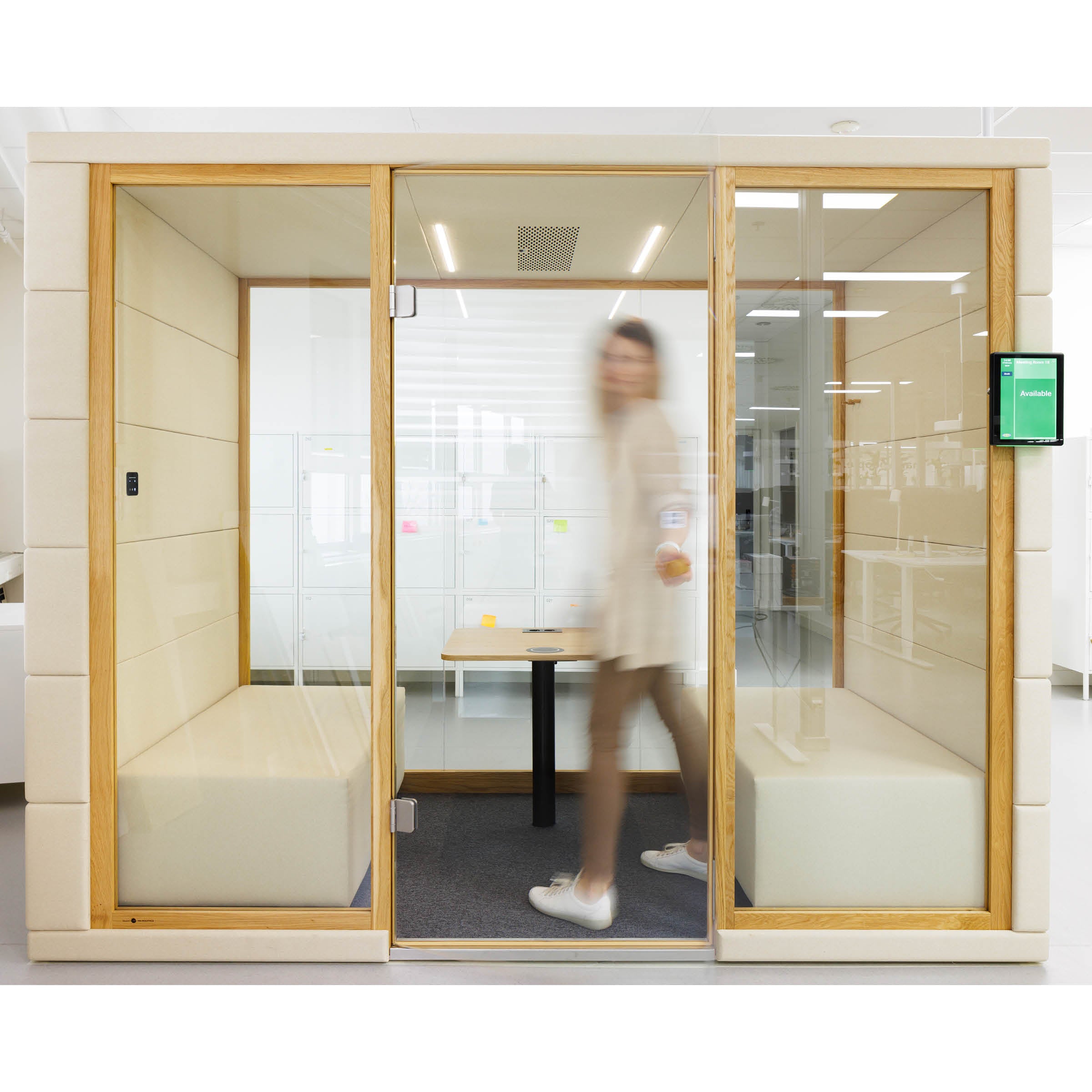 A woman strolls by an OFFICE POD QUADRIO 1 by SilentLab, featuring sleek wooden frames, a beige interior with cozy upholstered seating, and a small table. A digital sign is mounted outside the privacy-focused pod on the right.