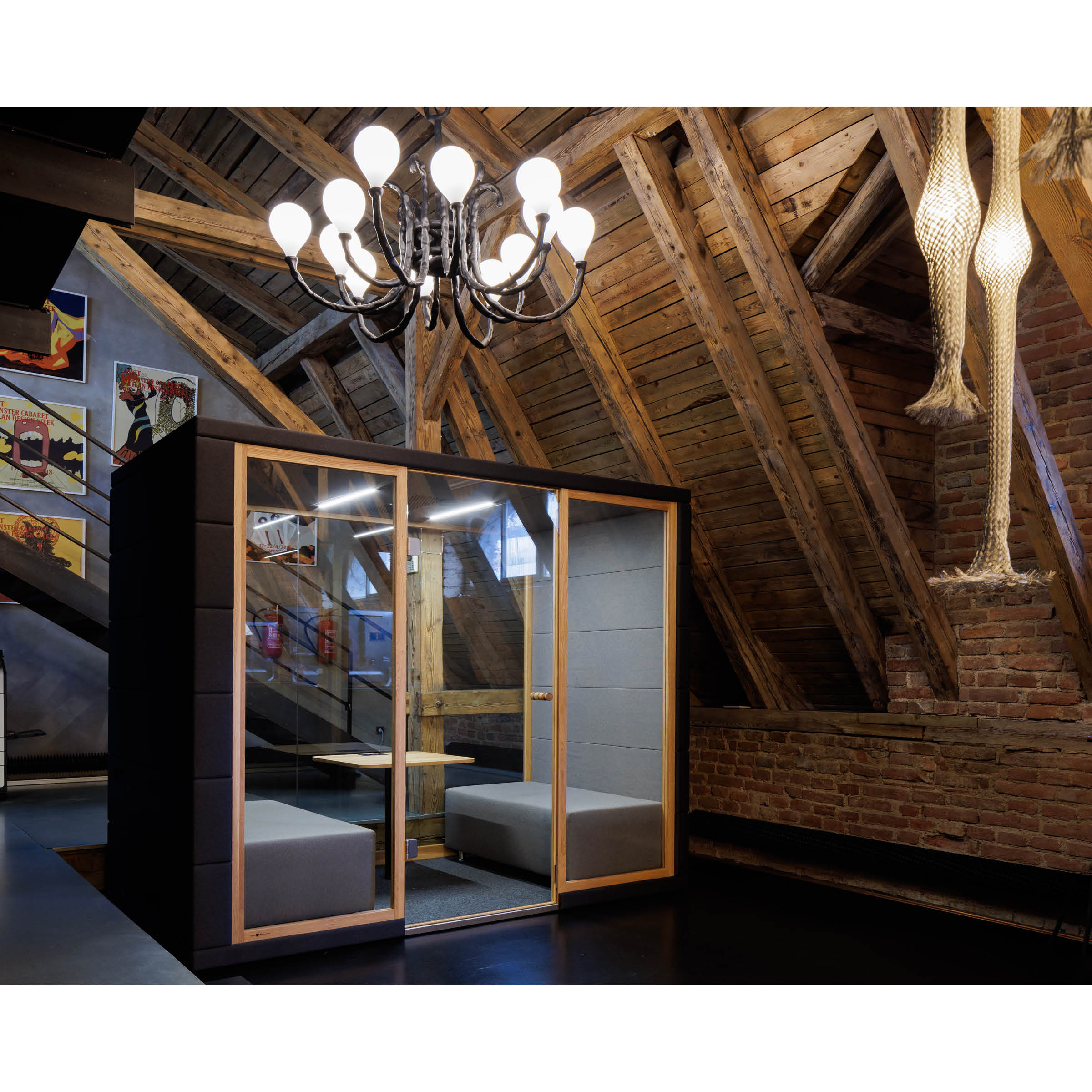The interior of this modern attic space boasts wooden beams, a sleek black and wood-accented phone booth, and the SilentLab OFFICE POD QUADRIO 1 for enhanced privacy. Illuminated by a chandelier, the area features brick walls, various artworks, and a unique fishnet-clad legs sculpture hanging from the ceiling.