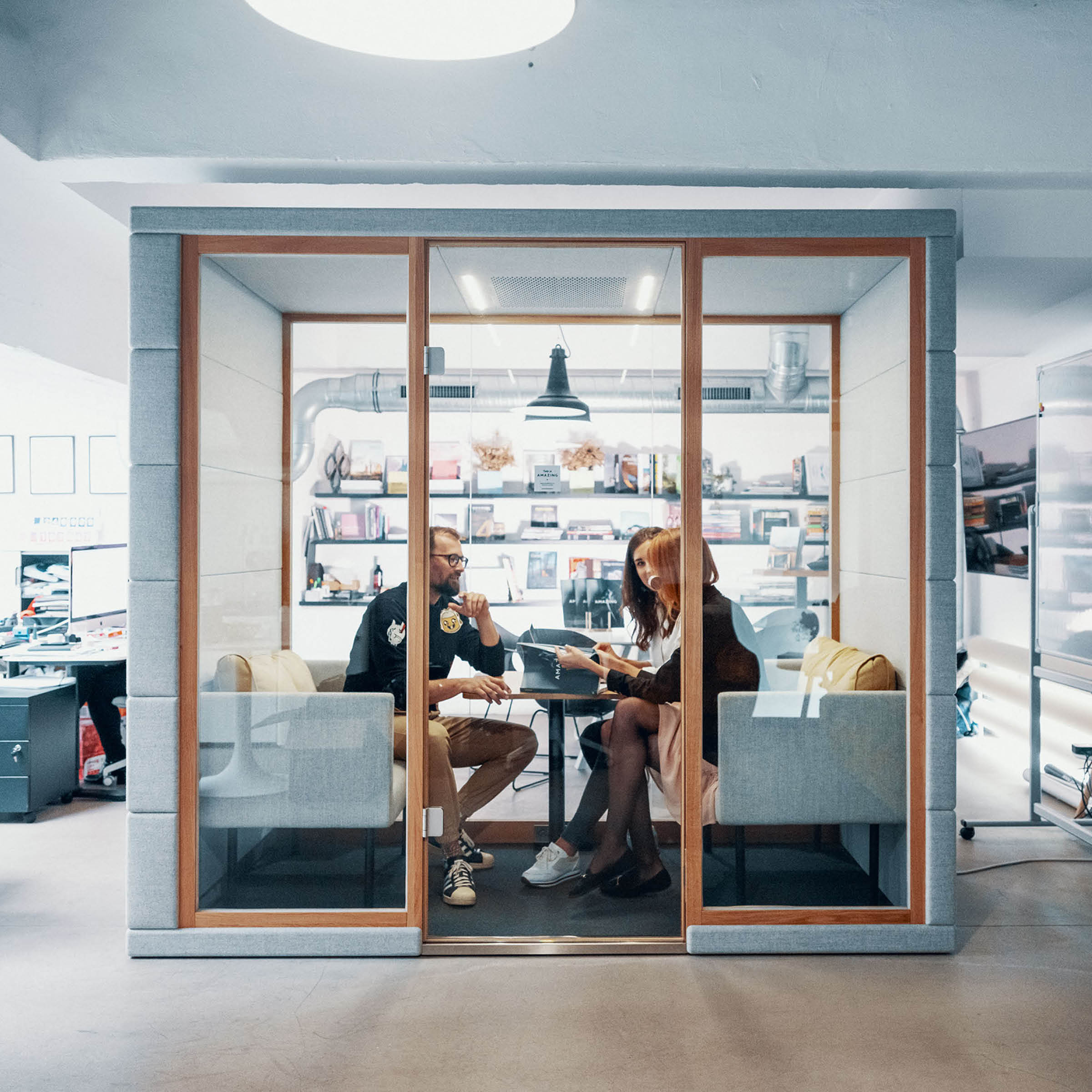 Inside a modern SilentLab OFFICE POD QUADRIO 1, two people converse at a small round table, surrounded by bookshelves. The bright and contemporary setting ensures privacy, with views of additional office areas through the glass walls.