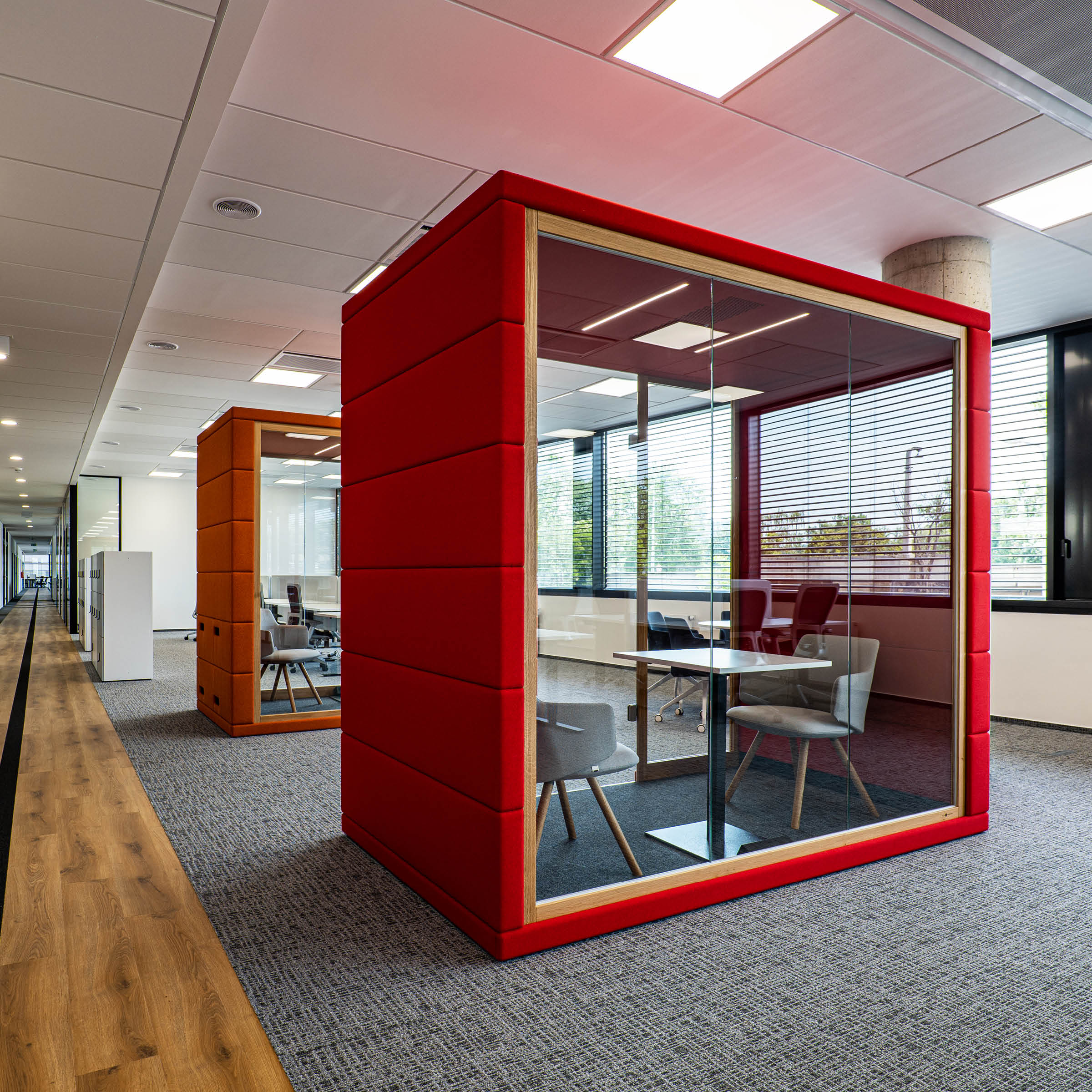 A modern office interior showcases two SilentLabs OFFICE POD QUADRIO 1 units with sleek, red glass walls. Each pod provides privacy with a table and chairs, complemented by gray carpet, wood flooring, and large windows that brighten the contemporary space with natural light.