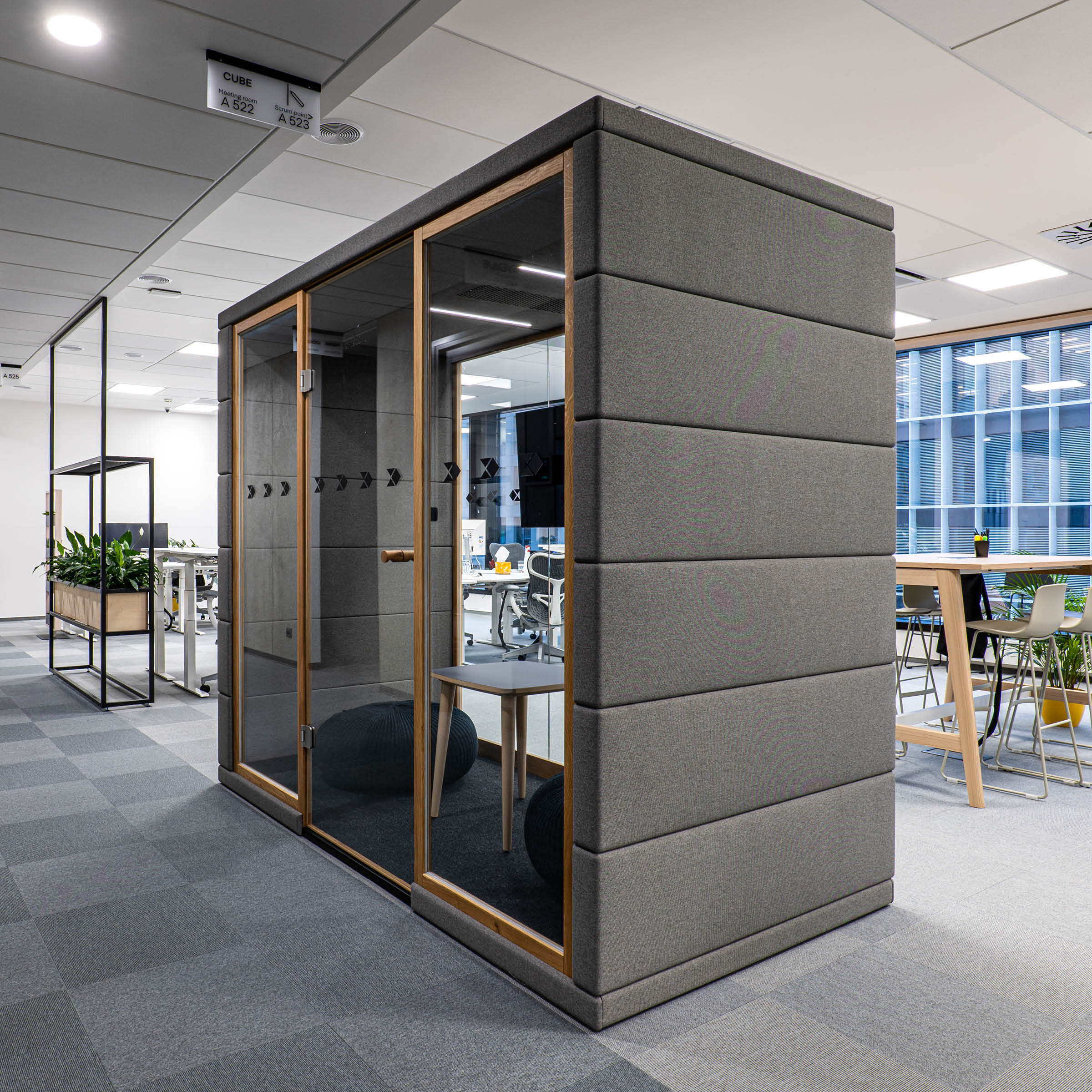 Discover the SilentLab OFFICE POD QUADRIO 1, a sleek modern office pod with glass-walled meeting space, ideal for privacy. It features padded gray panels and wooden accents. Inside are chairs and a small table, complemented by an open office area, desks, and greenery for a welcoming environment.