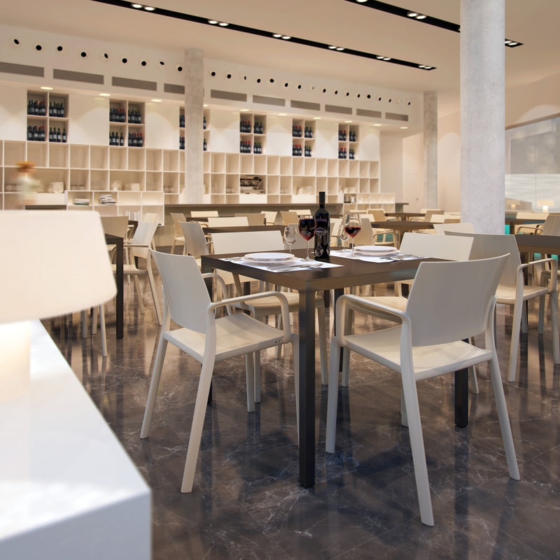 The modern restaurant interior boasts sleek white chairs with powder-coated aluminum frames and Resols Barcino Table set on a reflective marble floor, surrounded by wine shelves. Two wine glasses and a bottle adorn the table, complemented by white napkins. Bright lighting enhances the open ambiance.