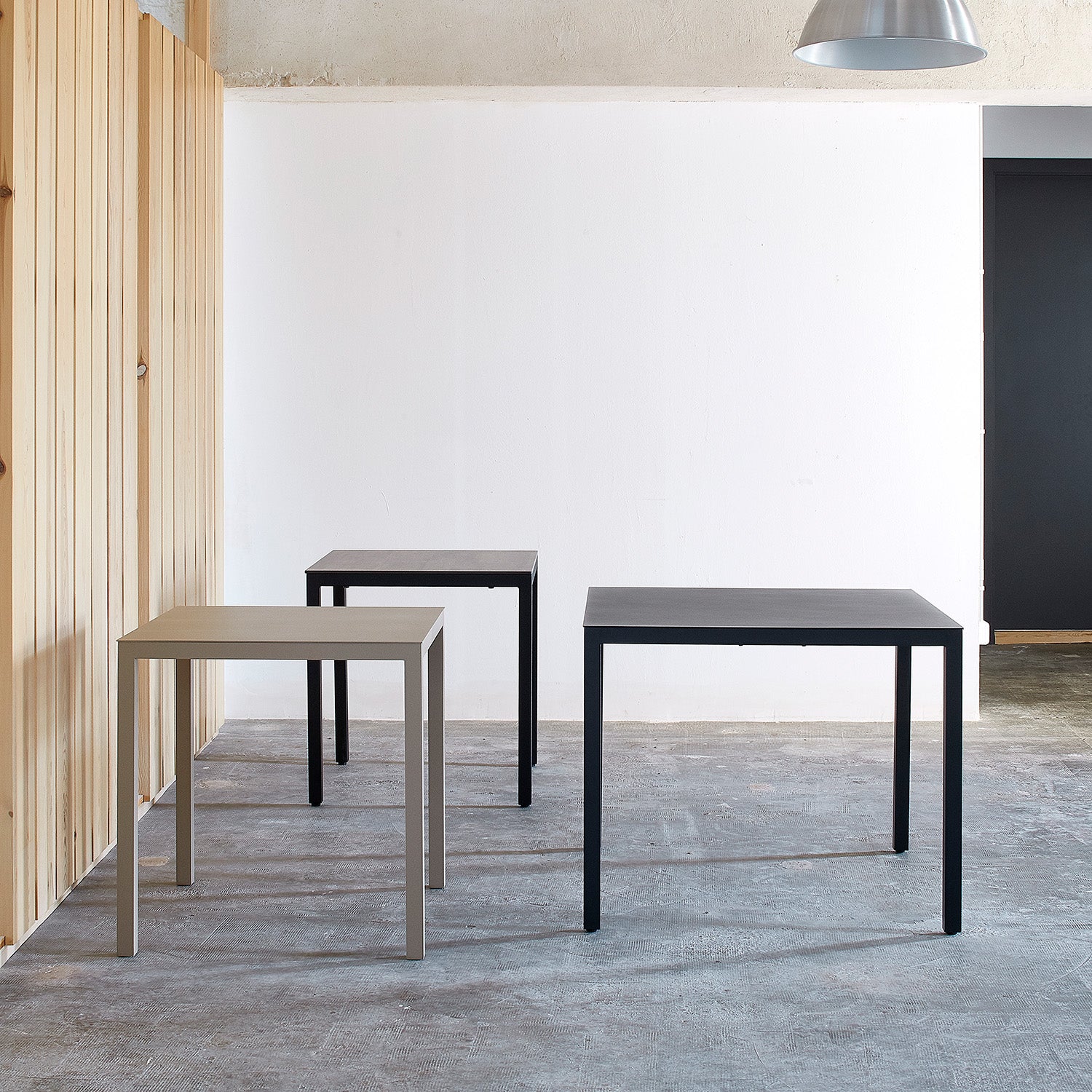 This minimalist room showcases three Barcino Tables by Resol, featuring square powder-coated aluminum frames in black and gray. Positioned against a white wall and wooden panel on a gray concrete floor beneath a large pendant light, they embody innovative furniture solutions.