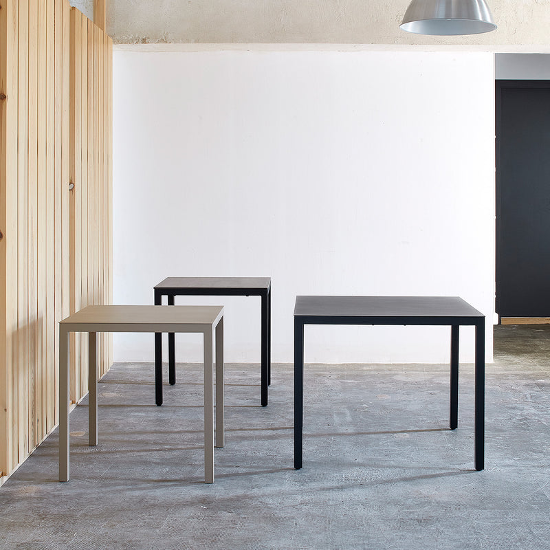 This minimalist room showcases three Barcino Tables by Resol, featuring square powder-coated aluminum frames in black and gray. Positioned against a white wall and wooden panel on a gray concrete floor beneath a large pendant light, they embody innovative furniture solutions.