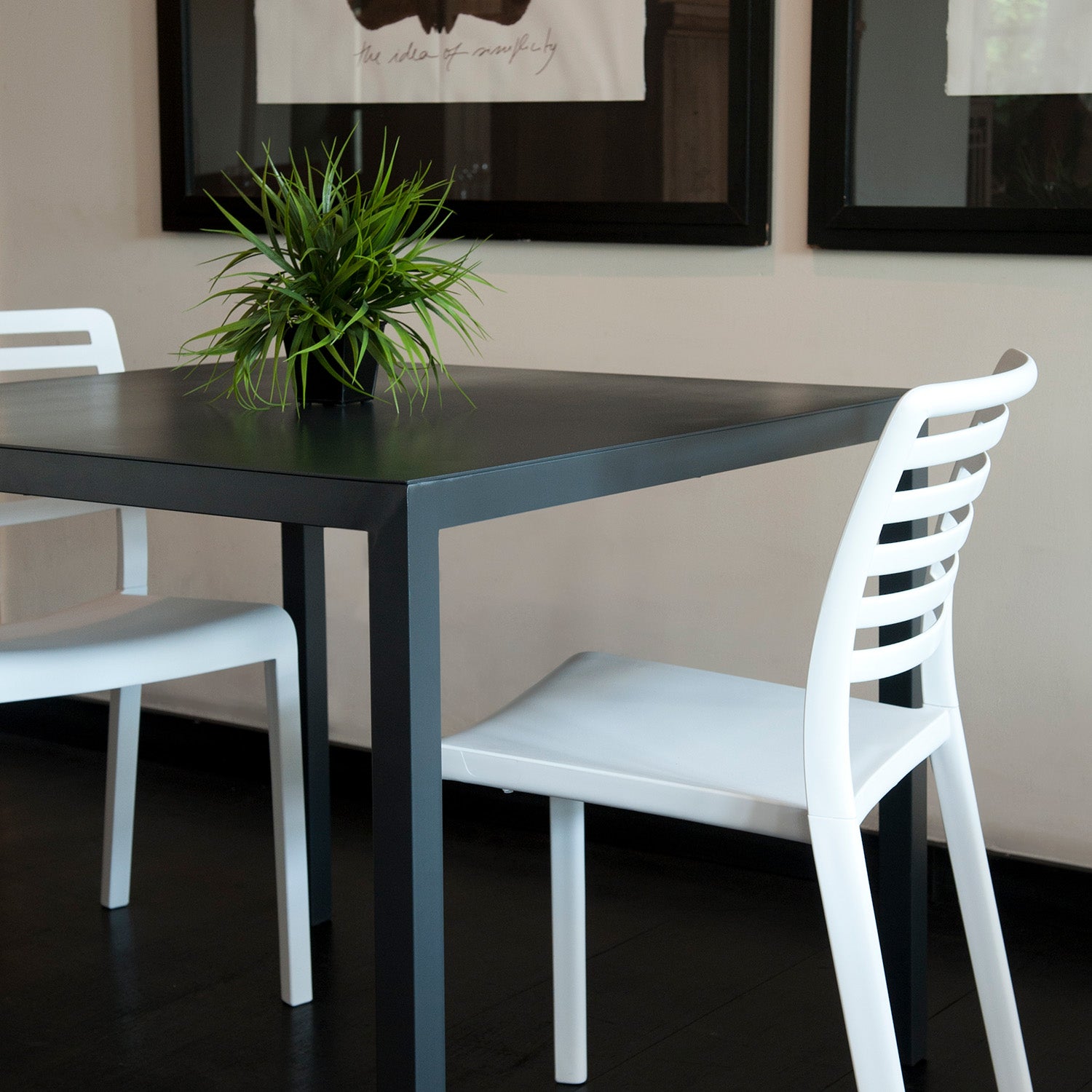 A modern dining setup showcases a black Barcino Table by Resol with a green plant centerpiece. Two white slatted-back chairs flank the table, surrounded by dark flooring and framed artwork on light-colored walls.