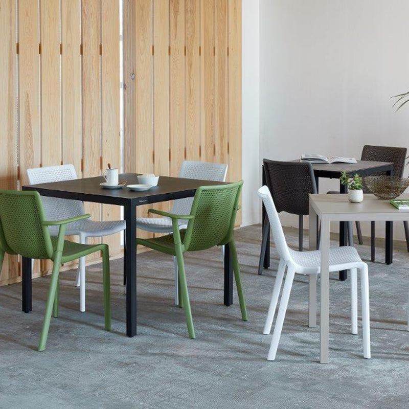 A modern café features light wood walls and several square Barcino tables by Resol. Each table, paired with colorful plastic chairs in green, white, and black, features a powder-coated aluminum frame. Cups, books, and a basket rest on the tables atop a gray textured carpet flooring.