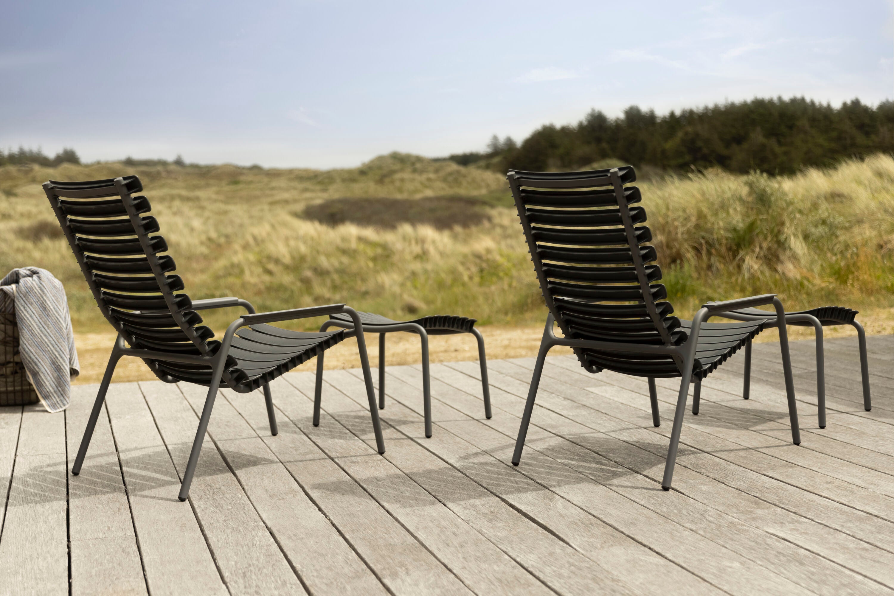 Two ReClips Footrests by Houe, along with matching modern black lounge chairs made from recycled plastic, are set on a wooden deck overlooking a grassy landscape under a clear sky. A striped towel on the deck adds coziness to this serene outdoor scene.