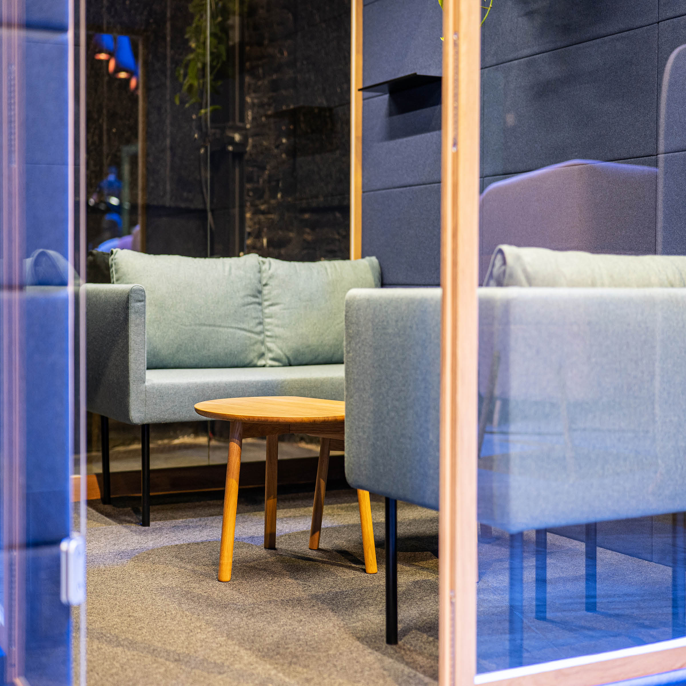 SilentLabs OFFICE POD REALM creates a cozy office meeting space with light green cushioned sofas, a round wooden coffee table, and a transparent glass door. Designed for comfort, it includes blue acoustic panels for workplace wellbeing and added privacy in this modern setting.