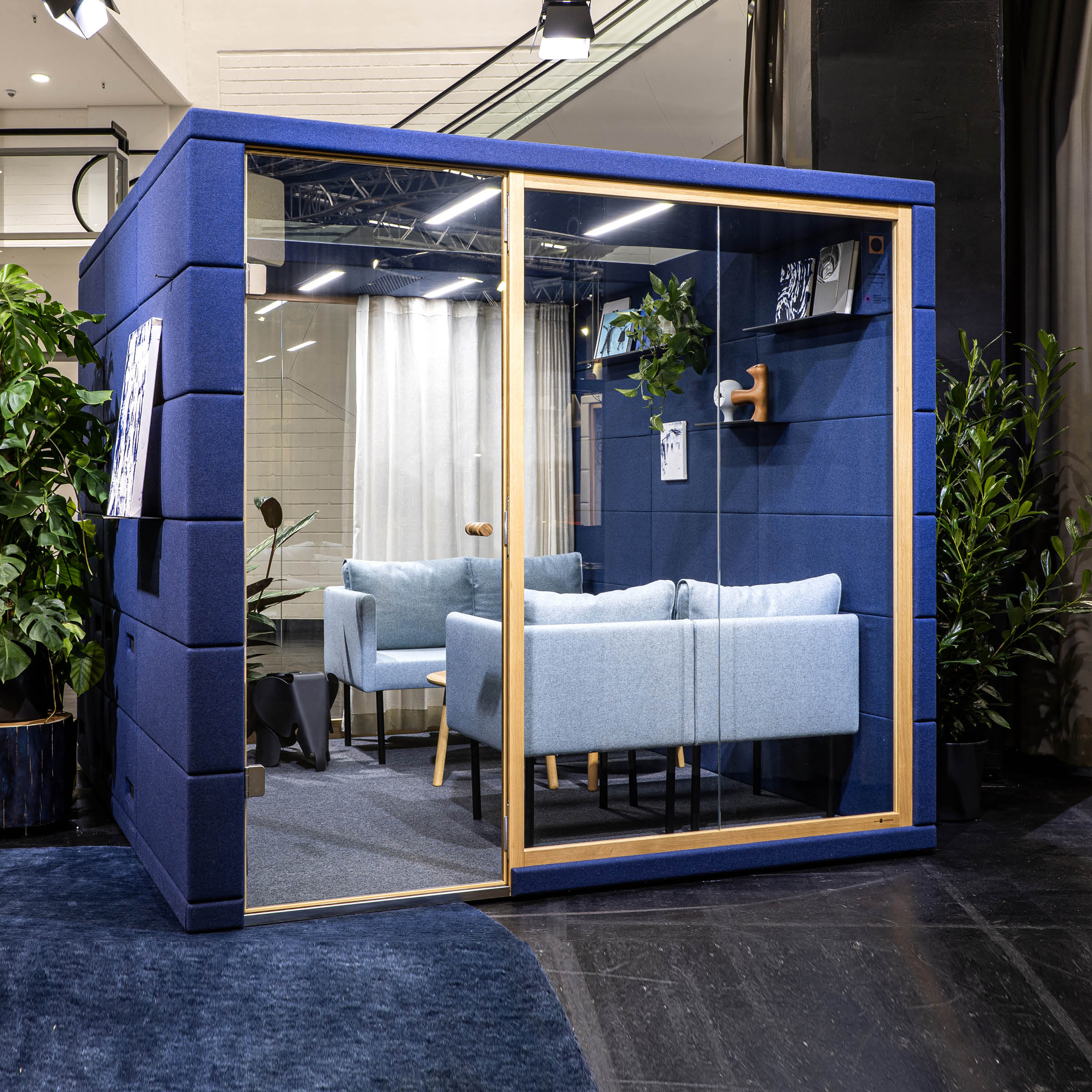 The SilentLab OFFICE POD REALM, a modern glass-walled acoustic office pod with a navy exterior, includes light blue sofas and a small round table. Its placed on dark carpet and features two potted plants and decorative items for enhanced workplace comfort.