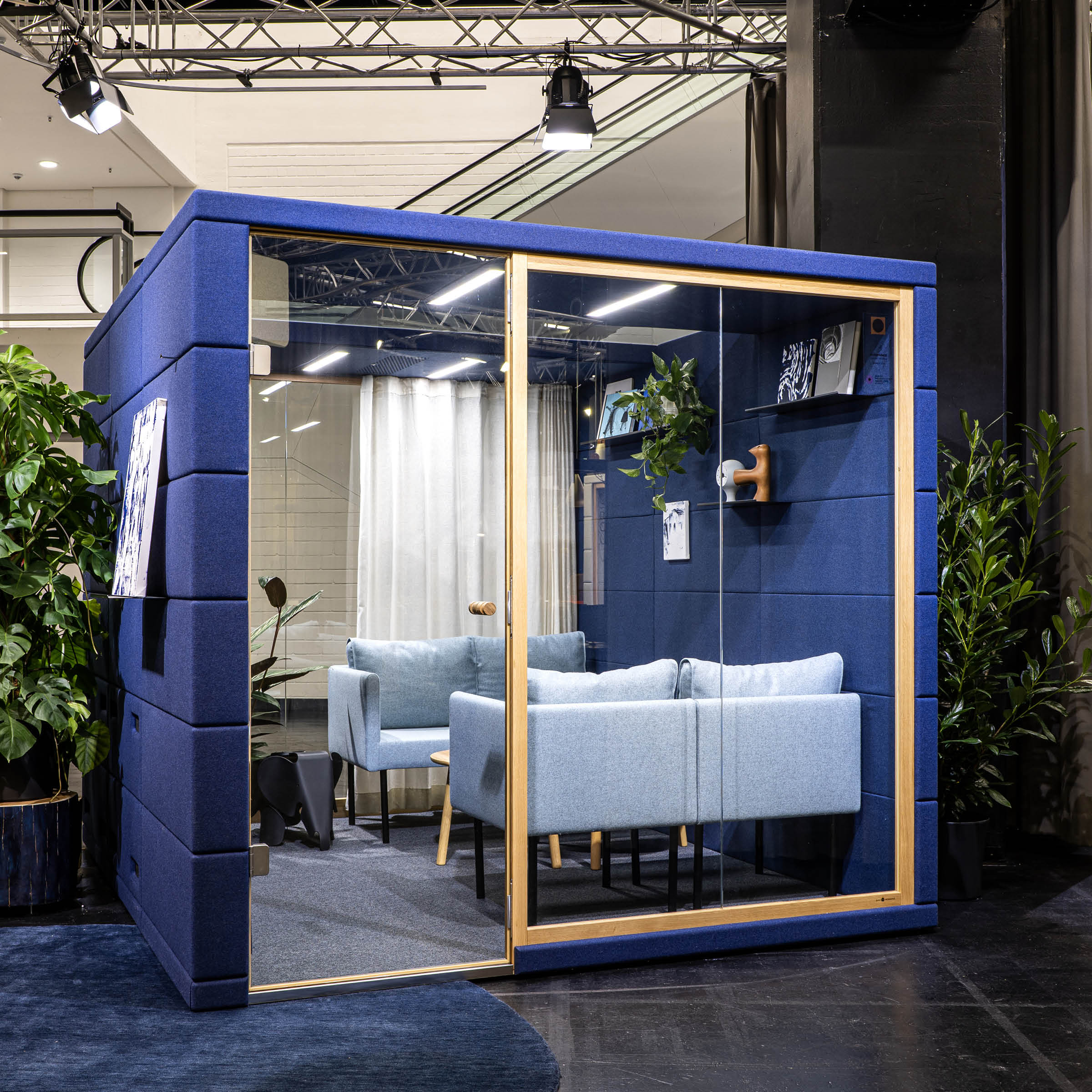 The OFFICE POD REALM by SilentLab is a modern acoustic pod with blue fabric walls and a glass door, featuring two light blue sofas and a small table. Surrounded by plants in an indoor setting with overhead lighting, it enhances workplace comfort and promotes wellbeing.