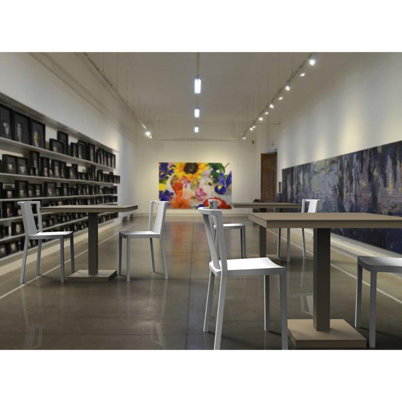 A modern art gallery interior with polished concrete floors showcases Resols Barcino Pie Central Square Tables and chairs. Walls adorned with colorful abstract artwork, shelves on the left display black cylinders, all brightly lit overhead in this sustainable production space.