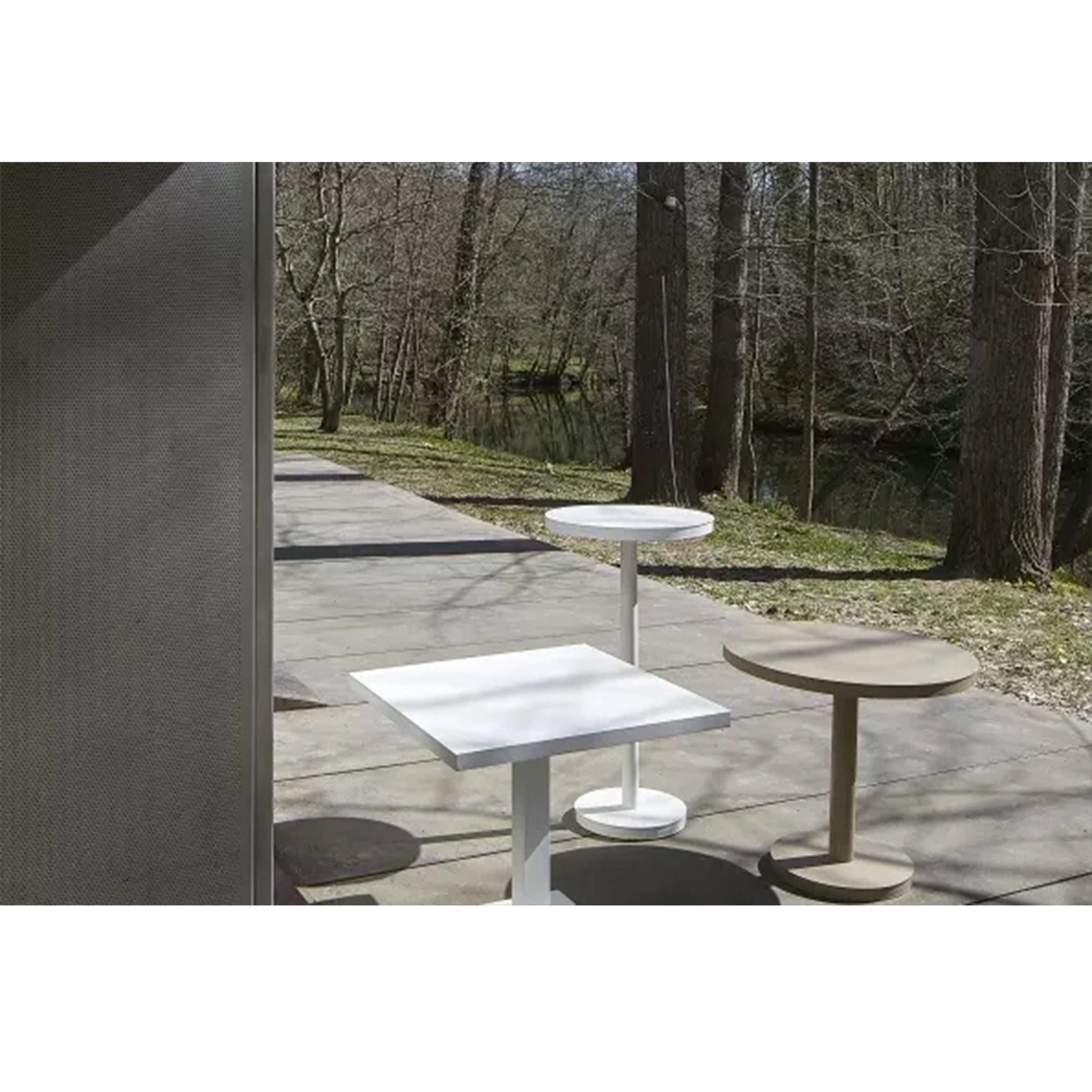 A Barcino Pie Central Square Table and two round Resol tables are set on a concrete patio. Surrounded by trees and soft sunlight, the natural setting and minimalistic, sustainably crafted furniture create a serene atmosphere.