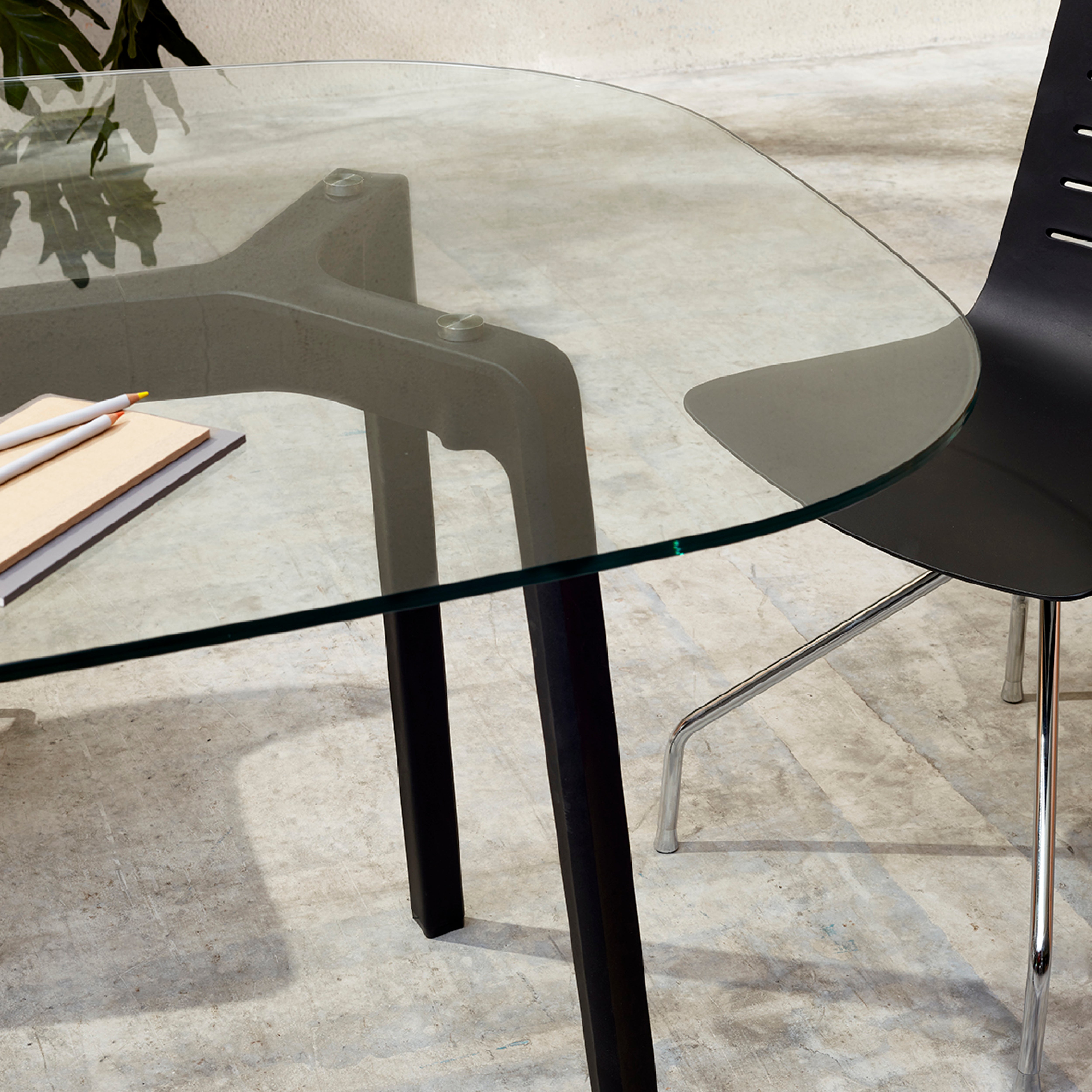 The Noren table by Resol, featuring a modern glass top and lacquered aluminum black legs, is paired with a black chair. A notebook and two pencils rest on the table, all set against a light surface that highlights the furnitures innovation and contemporary aesthetics through shadow play.