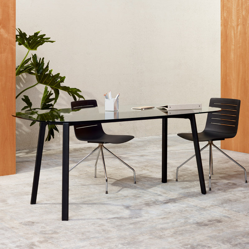 An office setup includes a Resol Noren glass-top table with black legs, two black chairs with metal legs, and a small plant in one corner. A white pen holder and a closed notebook rest on the table, all set against light-colored walls.