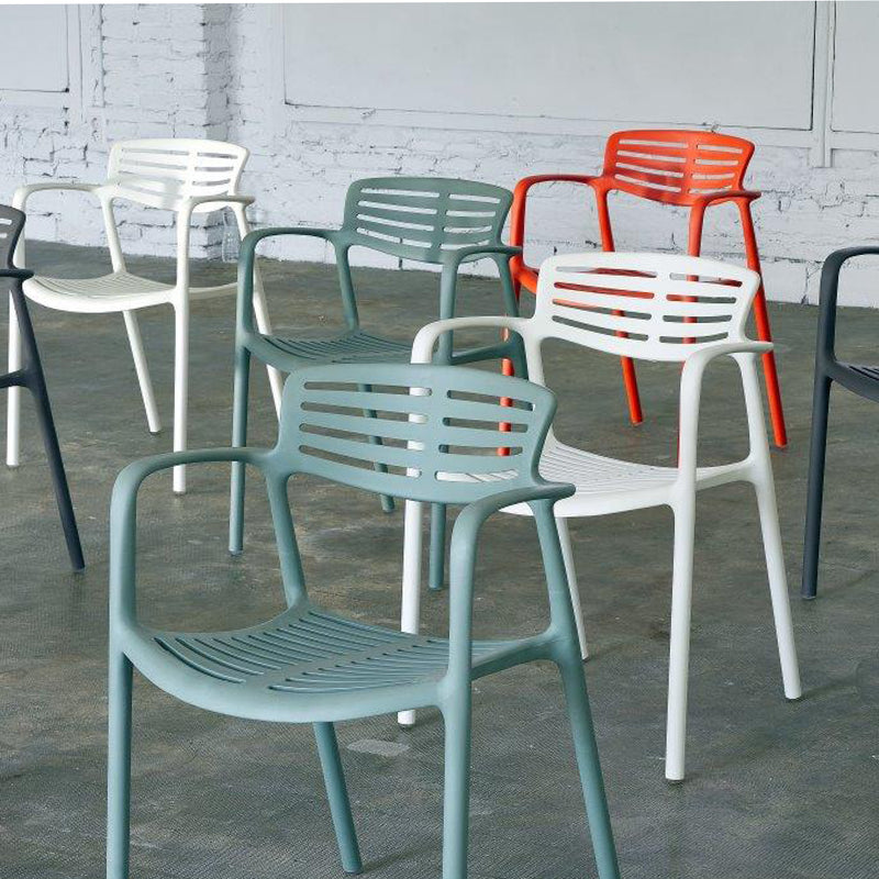A set of colorful Toledo Aire Armchairs by Resol, made using sustainable practices, is displayed on a concrete floor in front of a brick wall. These UV-protected, modern plastic chairs feature slatted backs and seats with armrests, available in white, gray, dark green, and orange.
