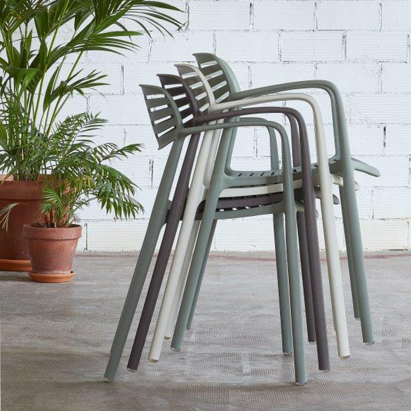 Four stackable Toledo Aire Armchairs by Resol, in shades of green and gray, are arranged side by side before a white brick wall. Crafted sustainably for an eco-friendly touch, theyre complemented by a leafy plant in a terracotta pot on the left.