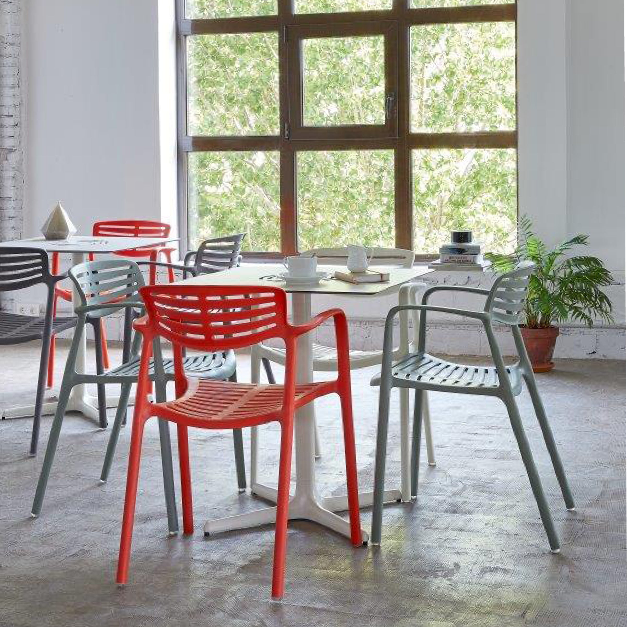 A bright room showcases innovative furniture solutions with modern Toledo Aire Armchairs by Resol in red and gray around round tables. A large window overlooks green trees, while a textured gray floor, a small plant, and a teapot set enhance the decor.