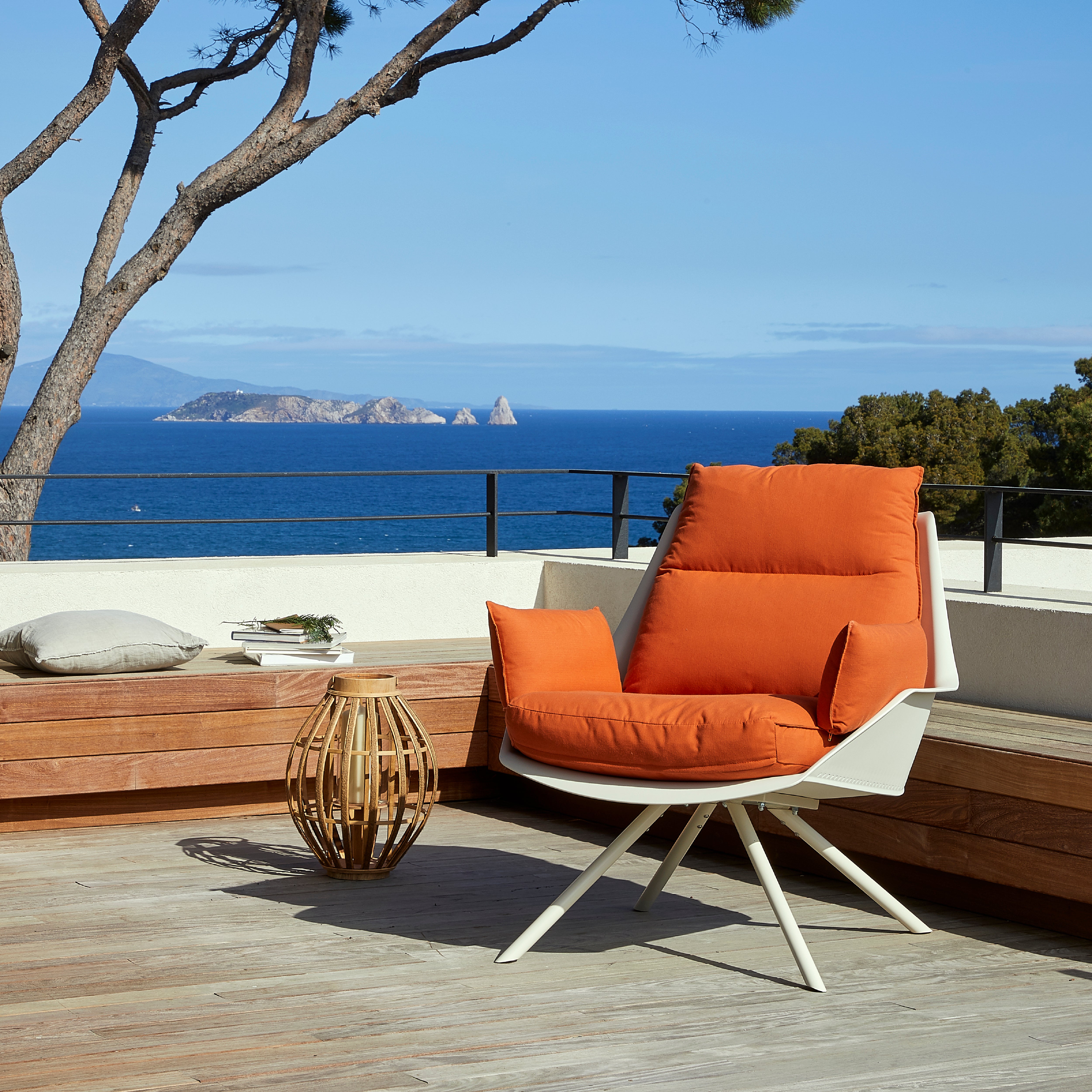 A chic patio overlooking the sea features a Resol Anou Metal Legs chair with an orange removable cushion and a polypropylene frame. The wooden deck is encircled by minimalist benches and trees, offering a distant view of a rocky island under a clear blue sky.
