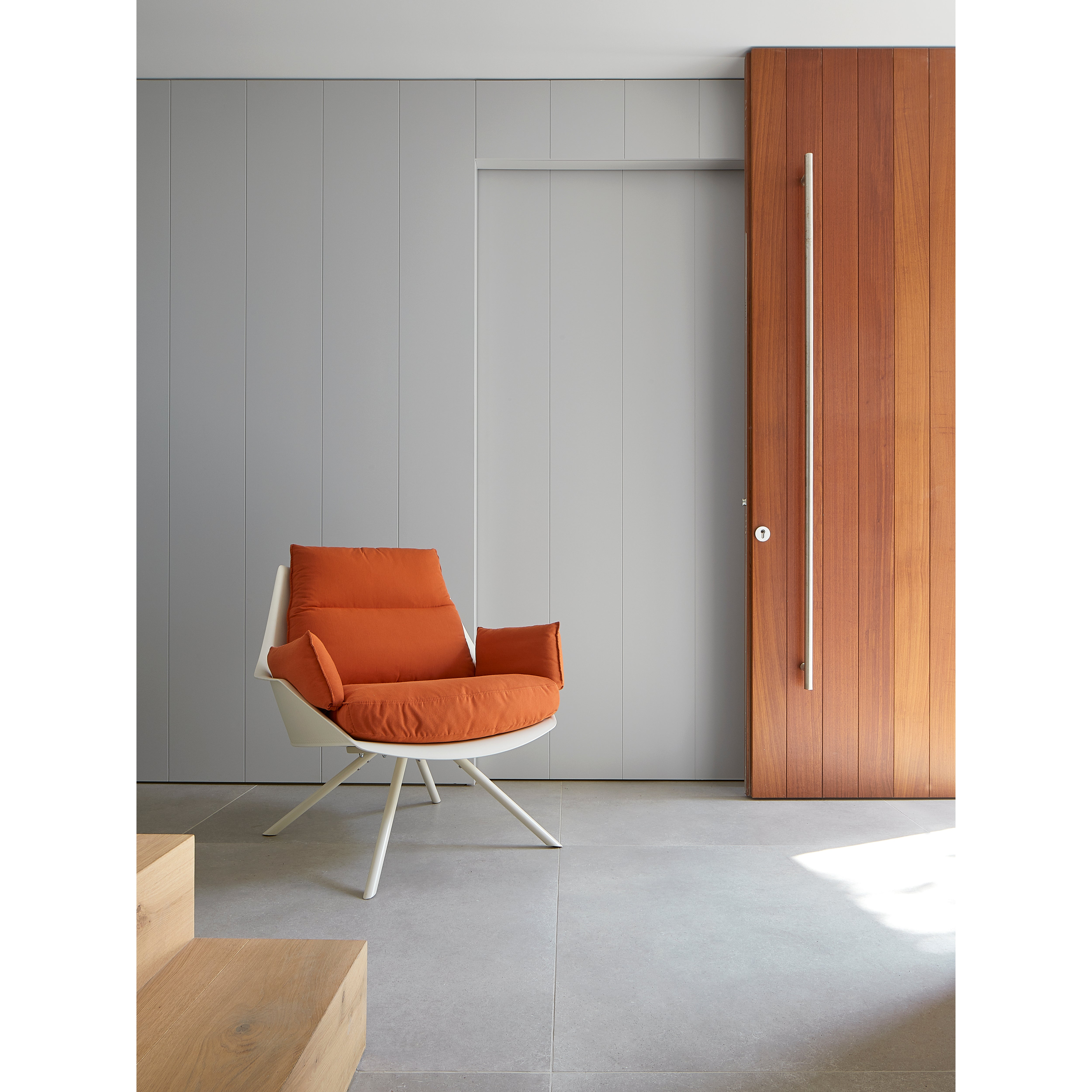 A modern room features a sleek Anou Metal Legs armchair by Resol in bright orange with a white metal base on a gray-tiled floor. Vertical gray panels and a wooden section with a handle enhance the minimalist style, all illuminated by natural light.