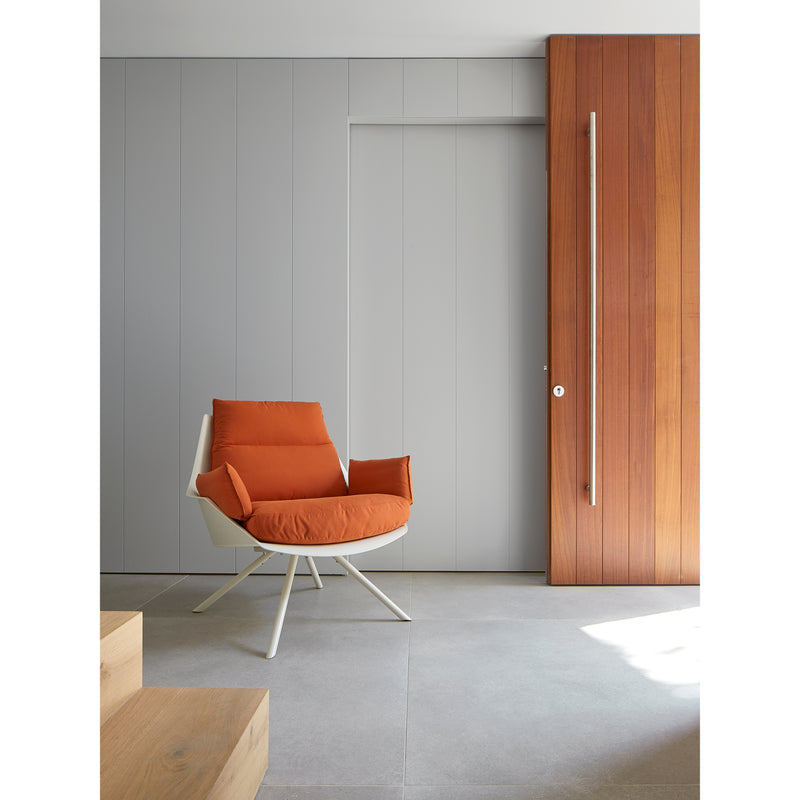 A modern room features a sleek Anou Metal Legs armchair by Resol in bright orange with a white metal base on a gray-tiled floor. Vertical gray panels and a wooden section with a handle enhance the minimalist style, all illuminated by natural light.
