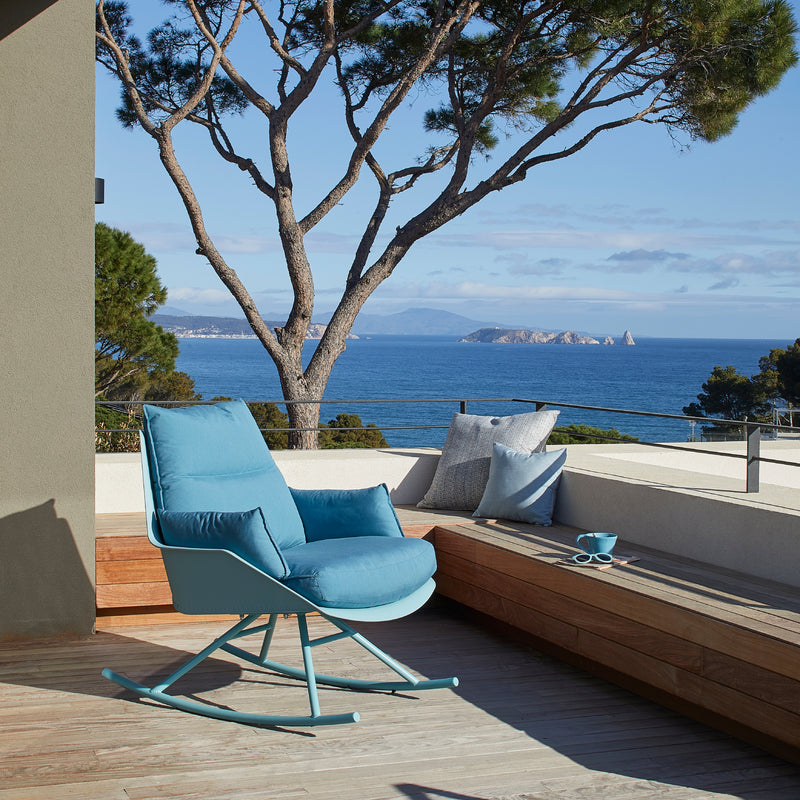 The terrace features a blue Anou Rocking Chair Legs by Resol with a polypropylene frame, perfectly paired with a pillow-adorned wooden bench. This serene spot offers sea and distant island views, framed by tall trees under a clear blue sky, blending indoor and outdoor elegance effortlessly.