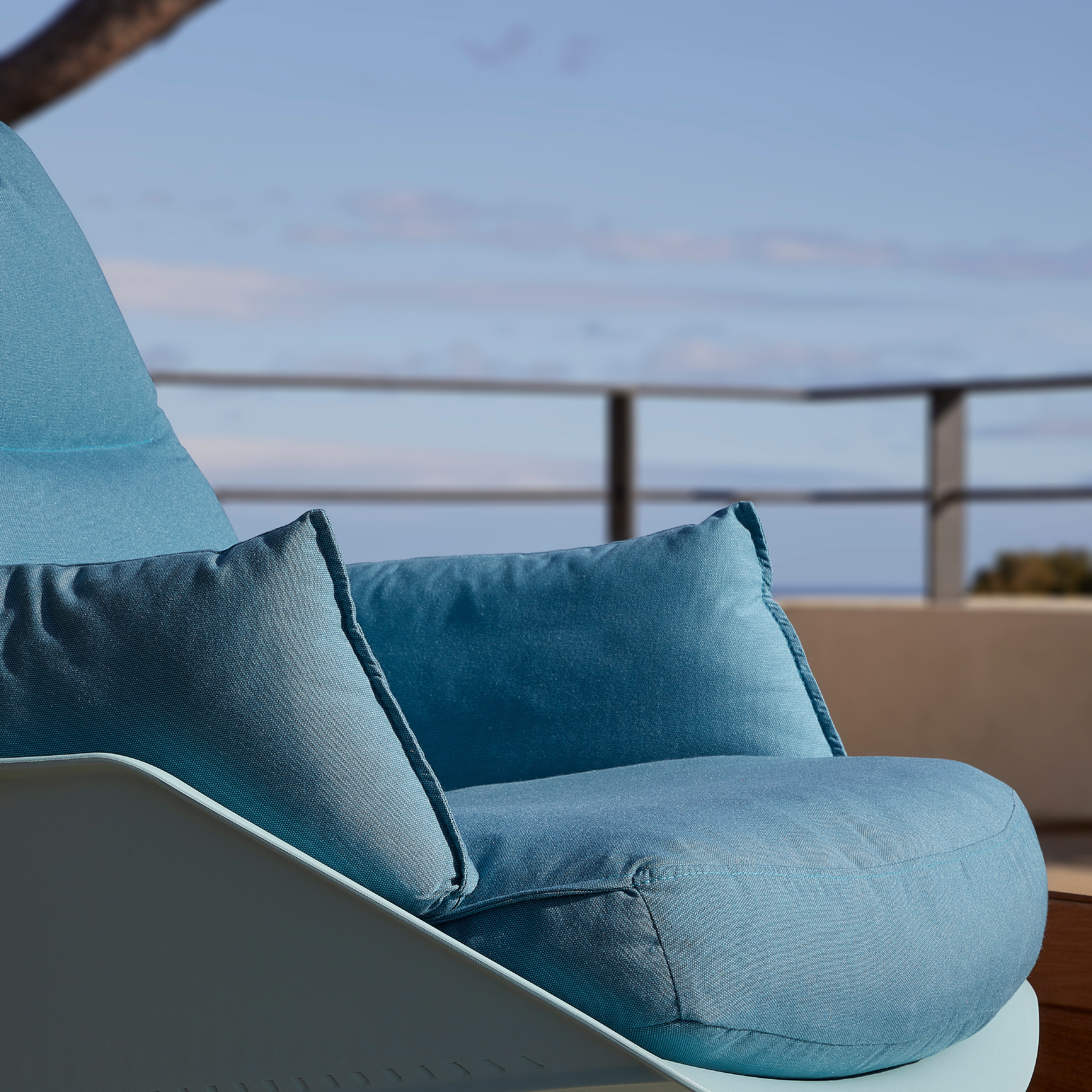 Close-up of a blue cushioned outdoor armchair, the Anou Rocking Chair Legs by Resol. Designed by Jorge Pensi, it features a sleek polypropylene frame on a sunny patio with a serene ocean view and softly focused distant railing and horizon.