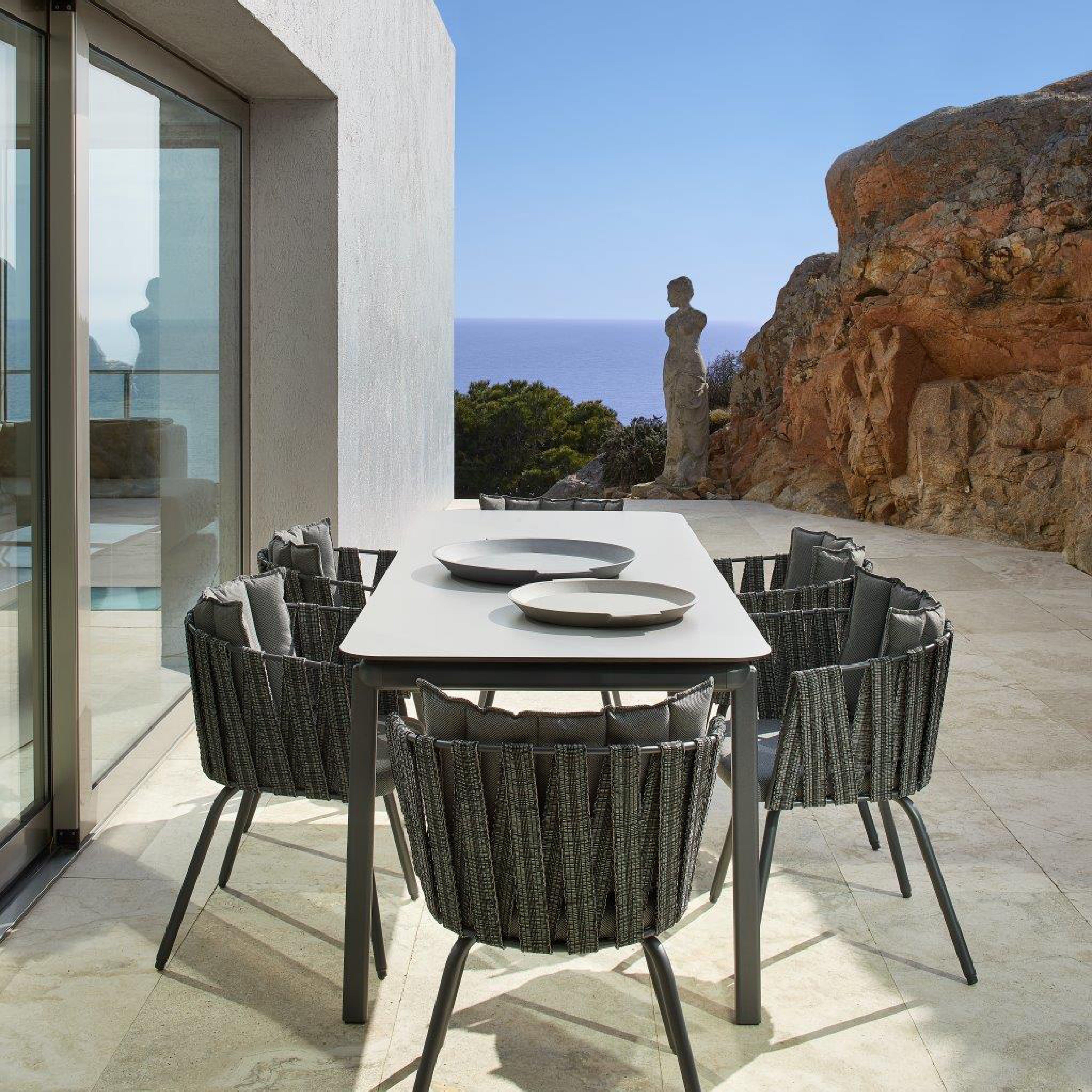 An outdoor dining area features the Resol Anthea Dining Table and six woven chairs on a stone terrace. Crafted sustainably, it overlooks rocky terrain and a stone sculpture with distant sea views under a clear sky. To the left are glass doors.