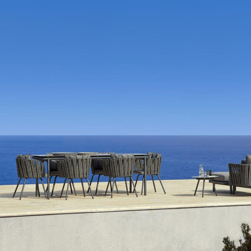 An outdoor patio with a Resol Anthea Dining Table and woven chairs overlooks a blue ocean and clear sky. Sparkling glassware adorns the table, while a sofa peeks from the right. This sustainable setting exudes a serene and open atmosphere.