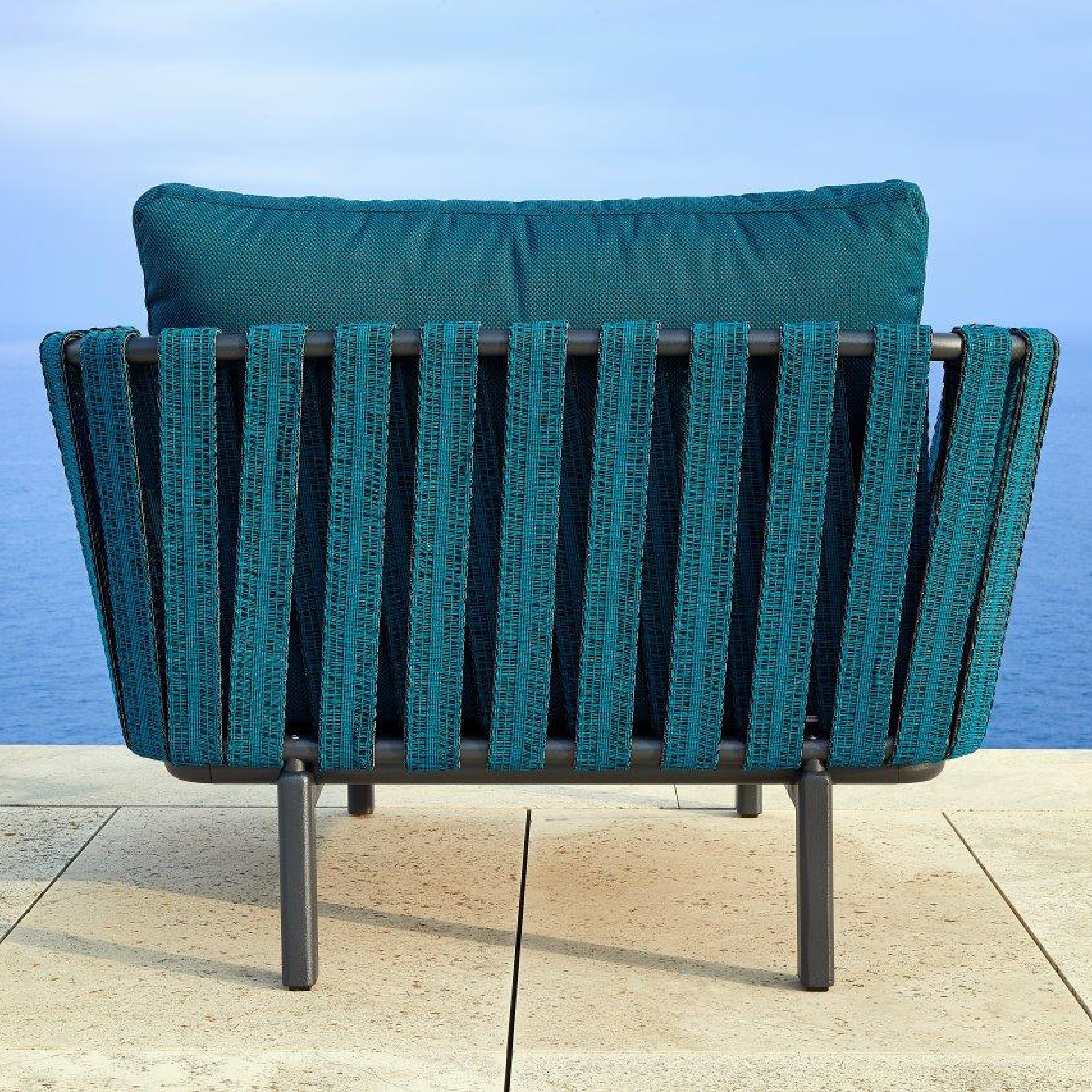 The Anthea Easy Chair by Resol, with a teal vertical slatted backrest and aluminum frame, is set on a beige stone terrace overlooking the blue ocean. The sky is bright and clear, enhancing the serene seaside atmosphere.