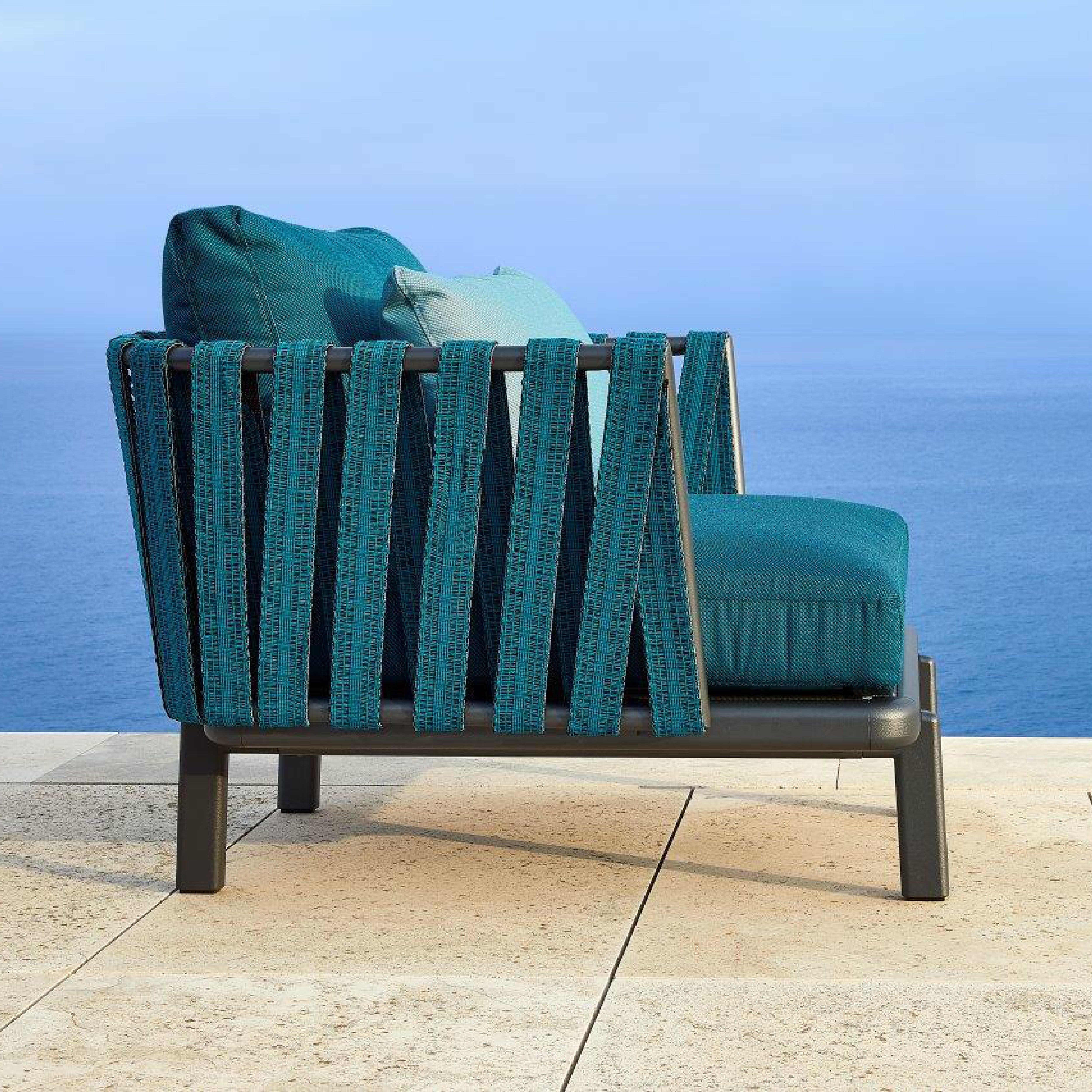 Side view of the Anthea Easy Chair by Resol with customizable teal cushions and a woven aluminum frame, set on a stone-tiled patio overlooking a calm blue ocean under a clear sky.