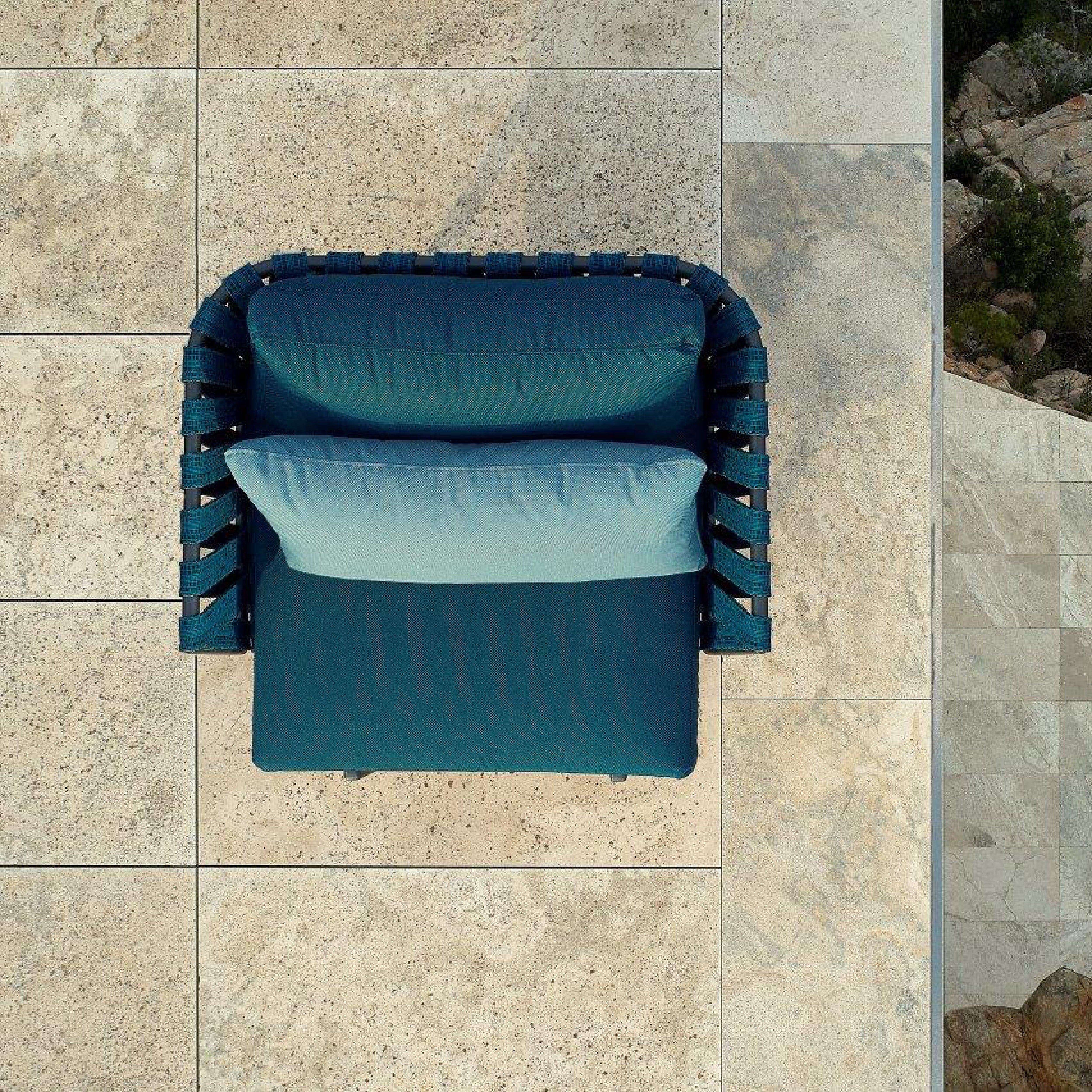 A top view of the Anthea Easy Chair by Resol, featuring a blue cushion on an aluminum frame, with woven backrests and armrests. A light blue rectangular pillow sits on the chair, which is placed on a beige stone patio, enhancing the stylish outdoor setting.