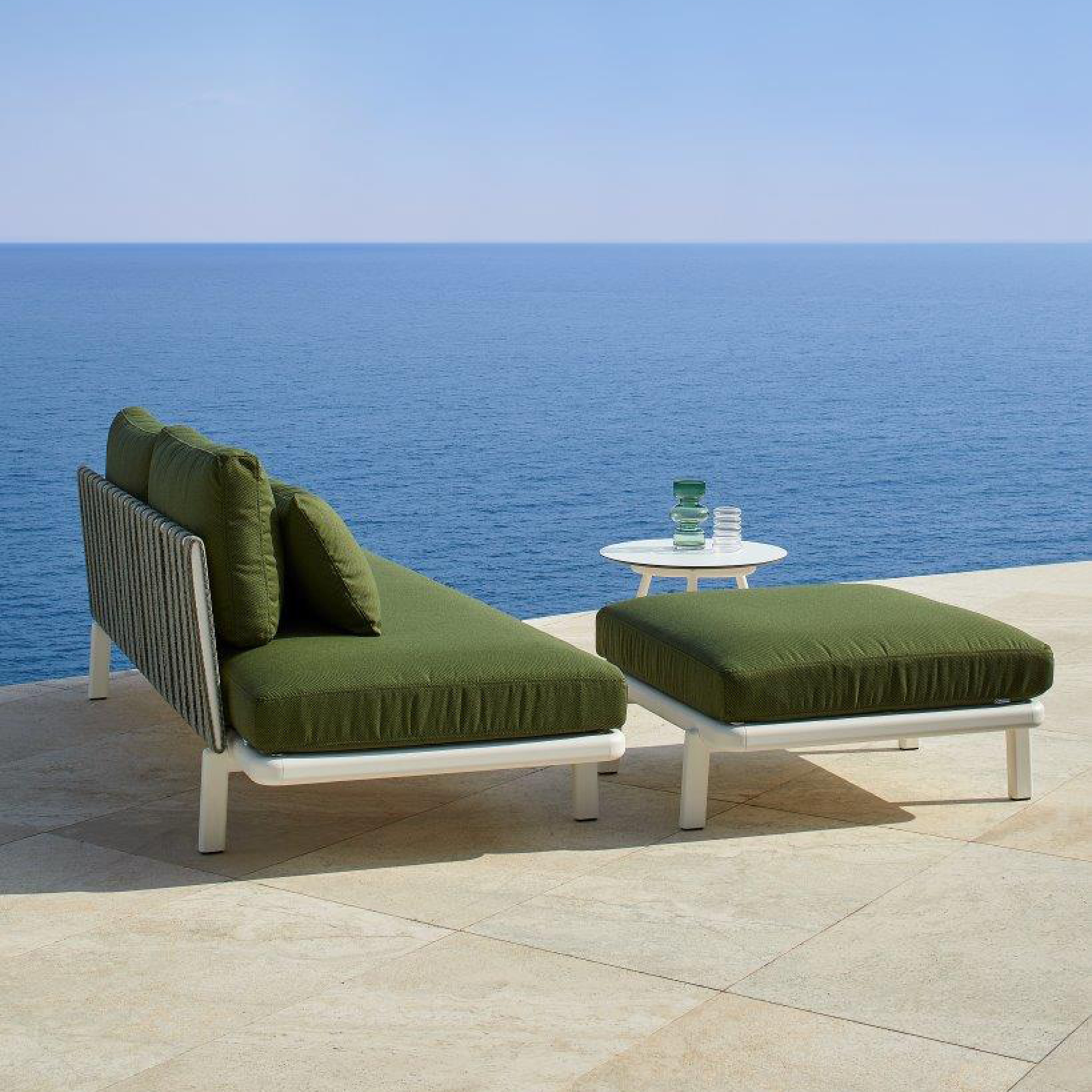 The Resol Anthea Ottoman set includes a green outdoor sofa with plush cushions, an ottoman, and a small round white table on a patio. The ensemble is framed in aluminum for outdoor durability, set against an ocean backdrop. Two glasses and a water bottle are placed on the table.