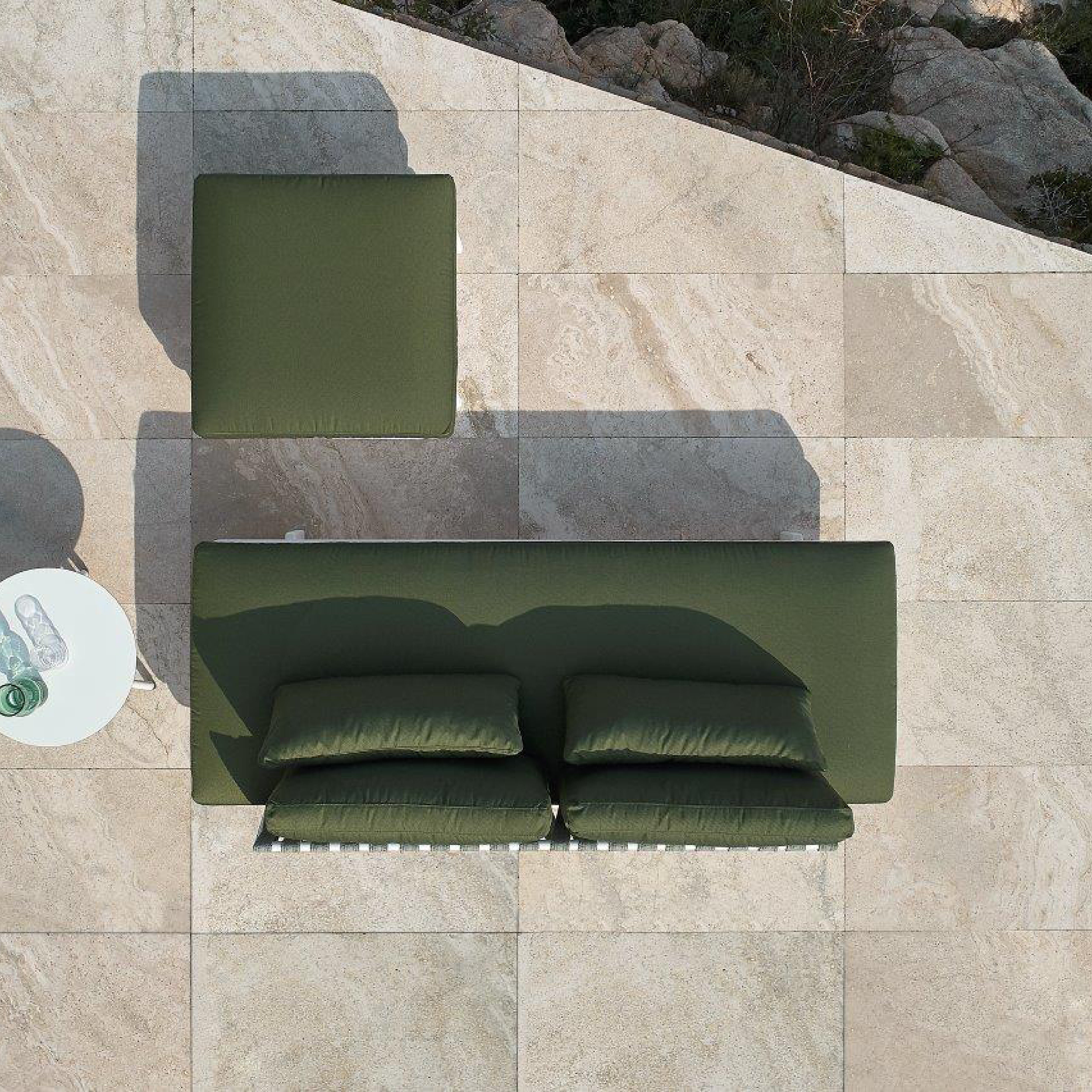 Top view of an outdoor patio setup featuring the Resol Anthea Ottoman with cushions on beige stone tiles. A small round white table holds a green bottle and a clear glass, all designed for outdoor use. Rocks and greenery enhance the serene background.