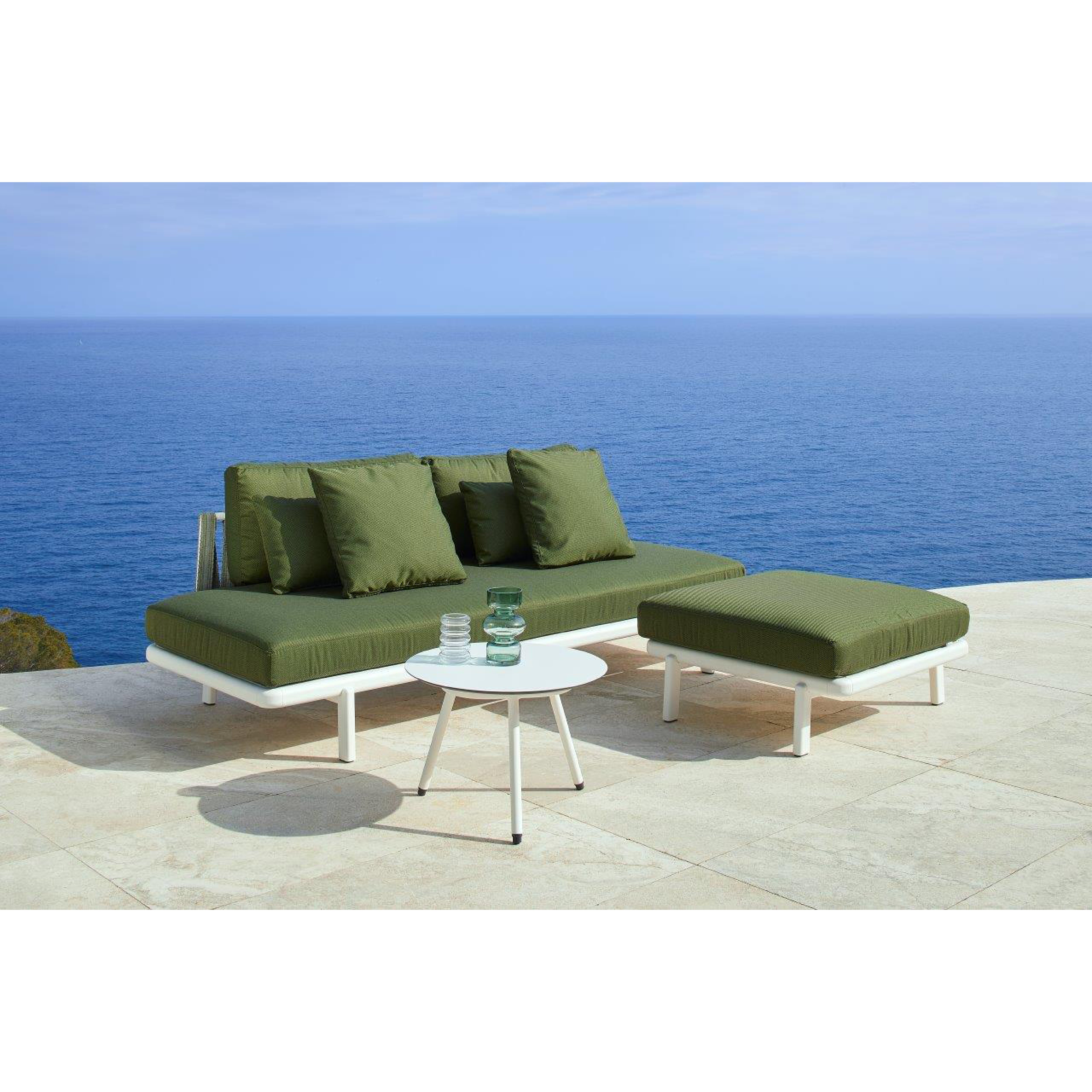 The Anthea Ottoman by Resol is a modern outdoor lounge set featuring green cushions and upholstery with a small white round table on a stone patio overlooking the ocean. Its durable aluminum frame ensures its ready for any season.