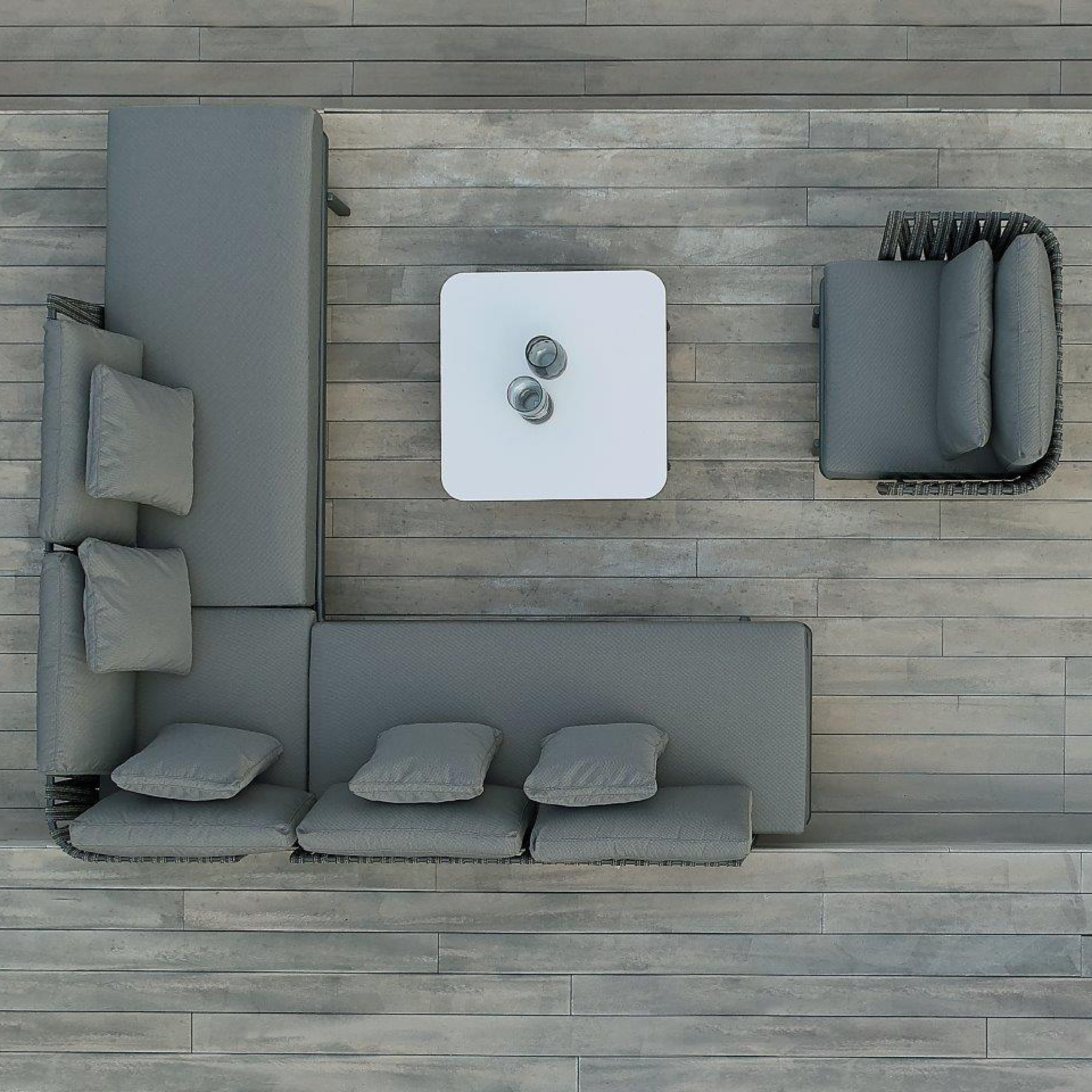 The Anthea Sofa System by Resol features a durable aluminum frame, five cushions, and an armchair positioned around a small white square table with two mugs. This elegant setup sits gracefully on a wooden deck as part of exquisite outdoor furniture seen from above.